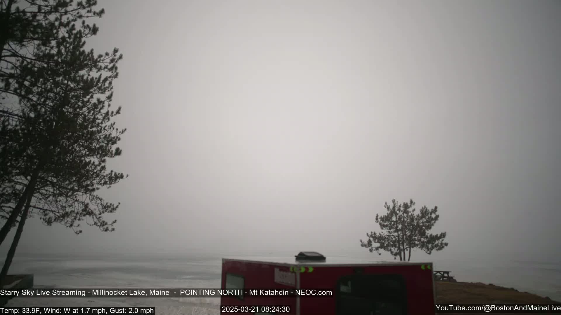 A foggy, grey scene shows a calm lake with a red vehicle and a few dark evergreen trees along the shoreline.