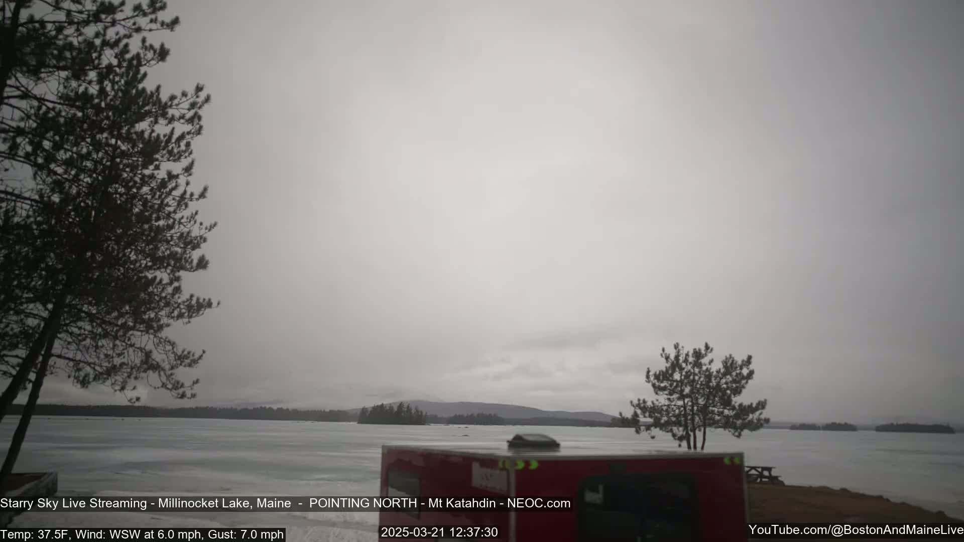 A mostly overcast, gray day shows a partially frozen lake with distant trees and hills under a flat, light gray sky.