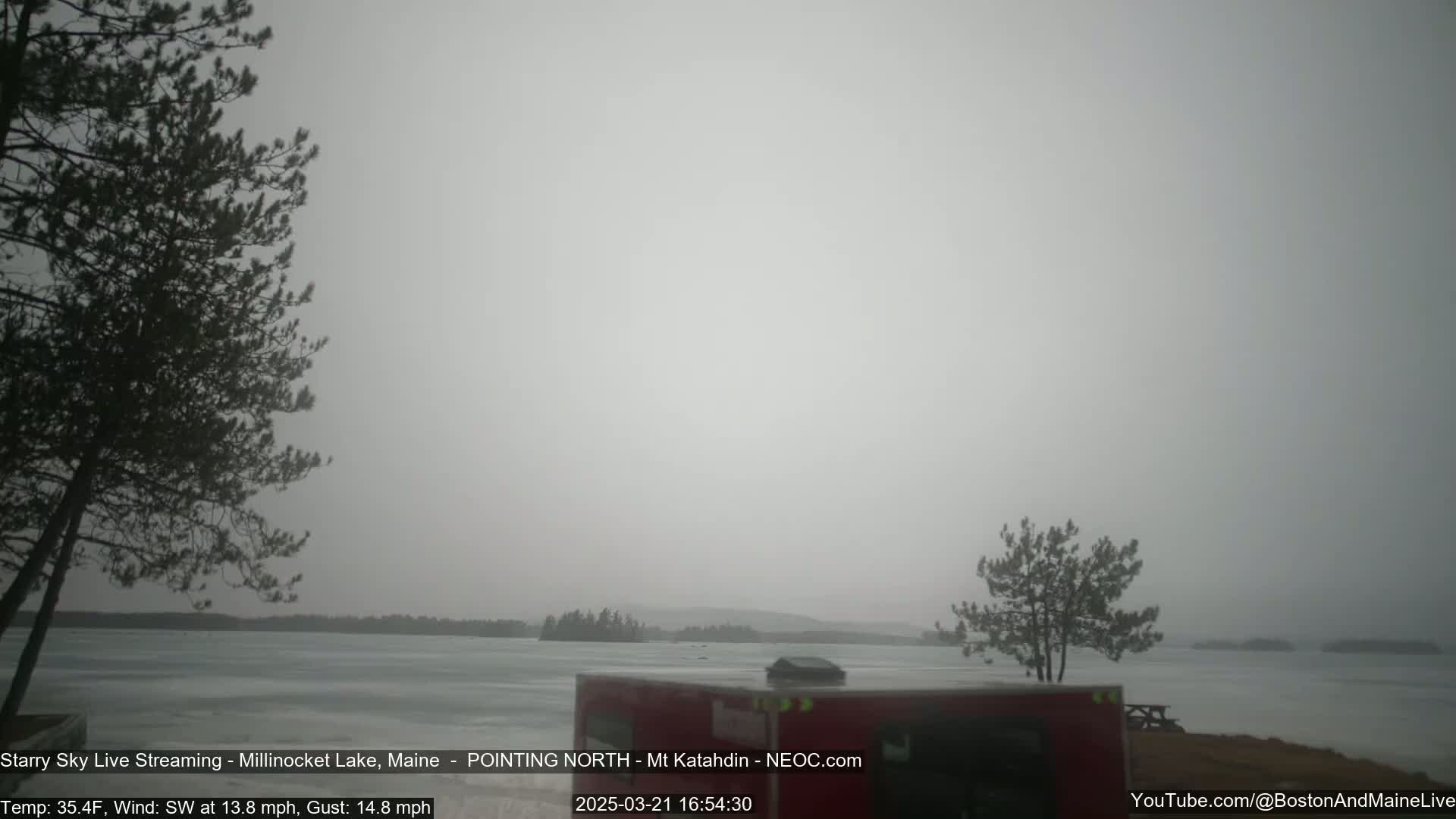 A mostly overcast, snowy scene shows a frozen lake with dark trees in the foreground and background, and a red camper in the bottom right corner.