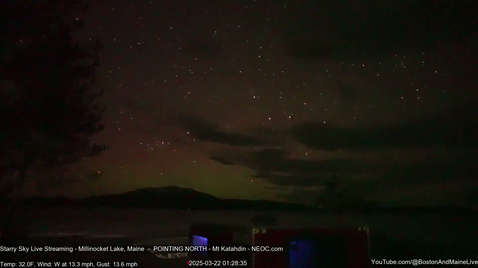 A nighttime view of a starry sky with a faint aurora borealis over a dark lake and silhouetted mountains, under a windy, 32°F temperature.