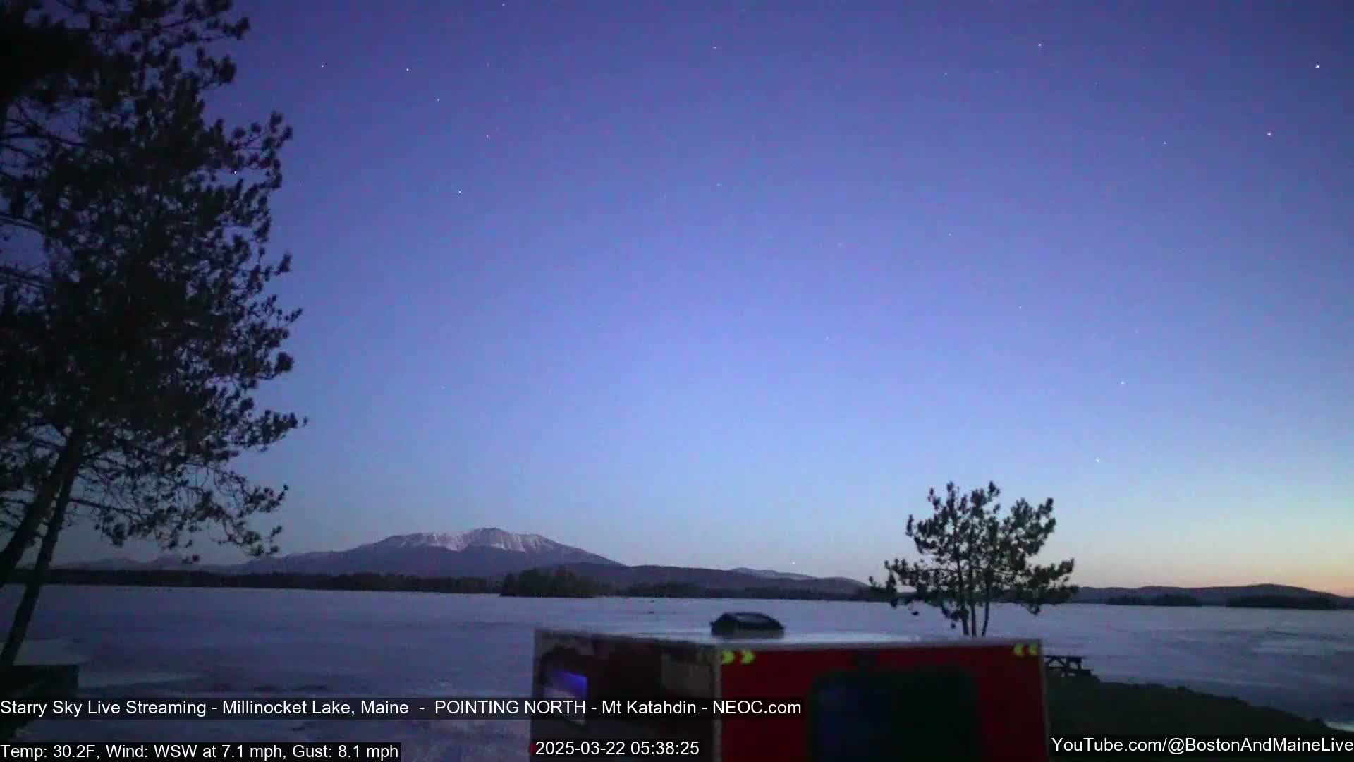 A starry, twilight sky over a frozen lake with snow-capped mountains in the distance and dark trees in the foreground, under a cold, windy WSW wind.