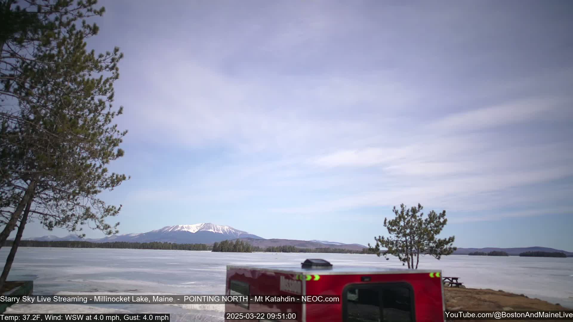 A partially frozen lake with a distant snow-capped mountain range under a mostly clear, light blue sky is viewed from behind a red camper van.