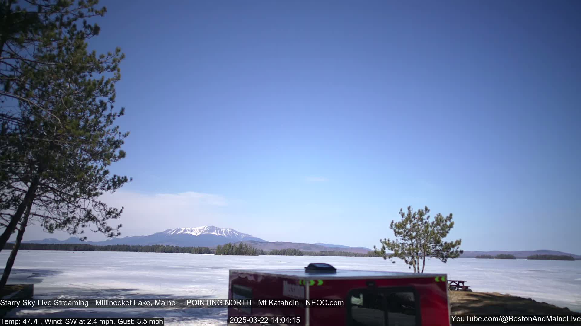 A mostly clear blue sky overlooks a frozen lake with a distant mountain range, and a red camper is partially visible in the foreground.