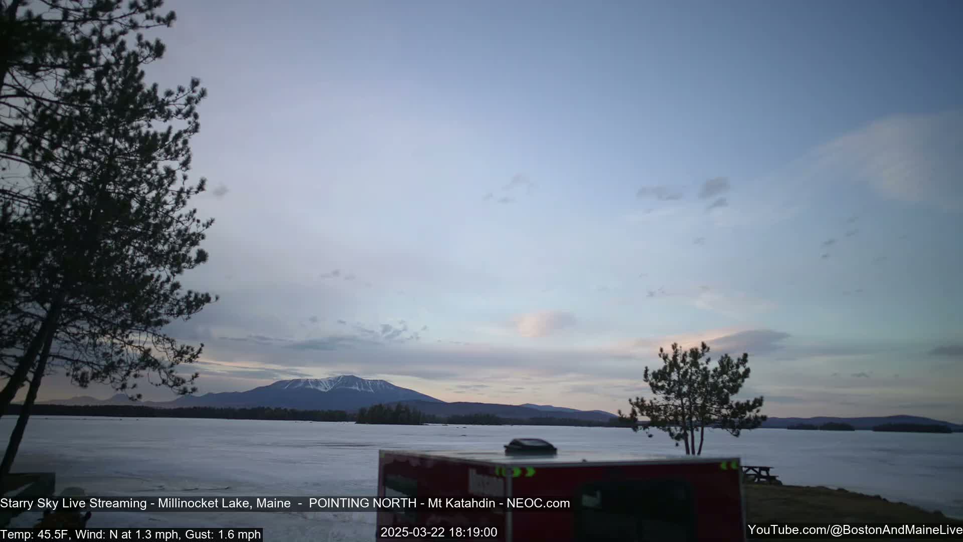 A partly cloudy twilight sky over a frozen lake with a mountain range in the distance, and a few dark evergreen trees in the foreground.