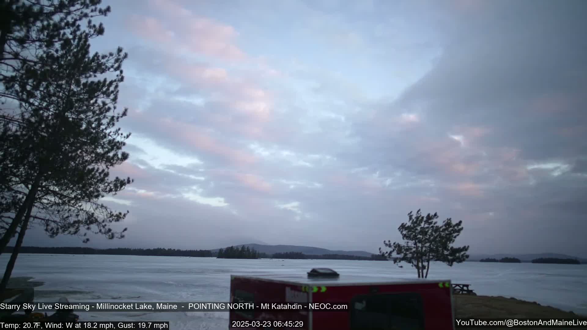 A partially frozen lake is viewed from a shoreline with a camper and trees under a cloudy, pale pink and gray sky with a brisk wind.