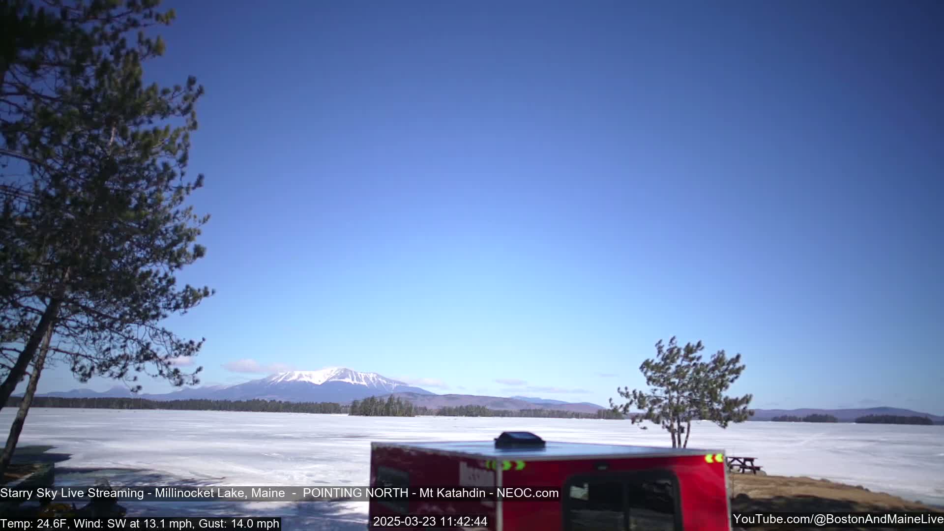 A red camper van is parked near a frozen lake under a clear blue sky, with a distant mountain range visible in the background.