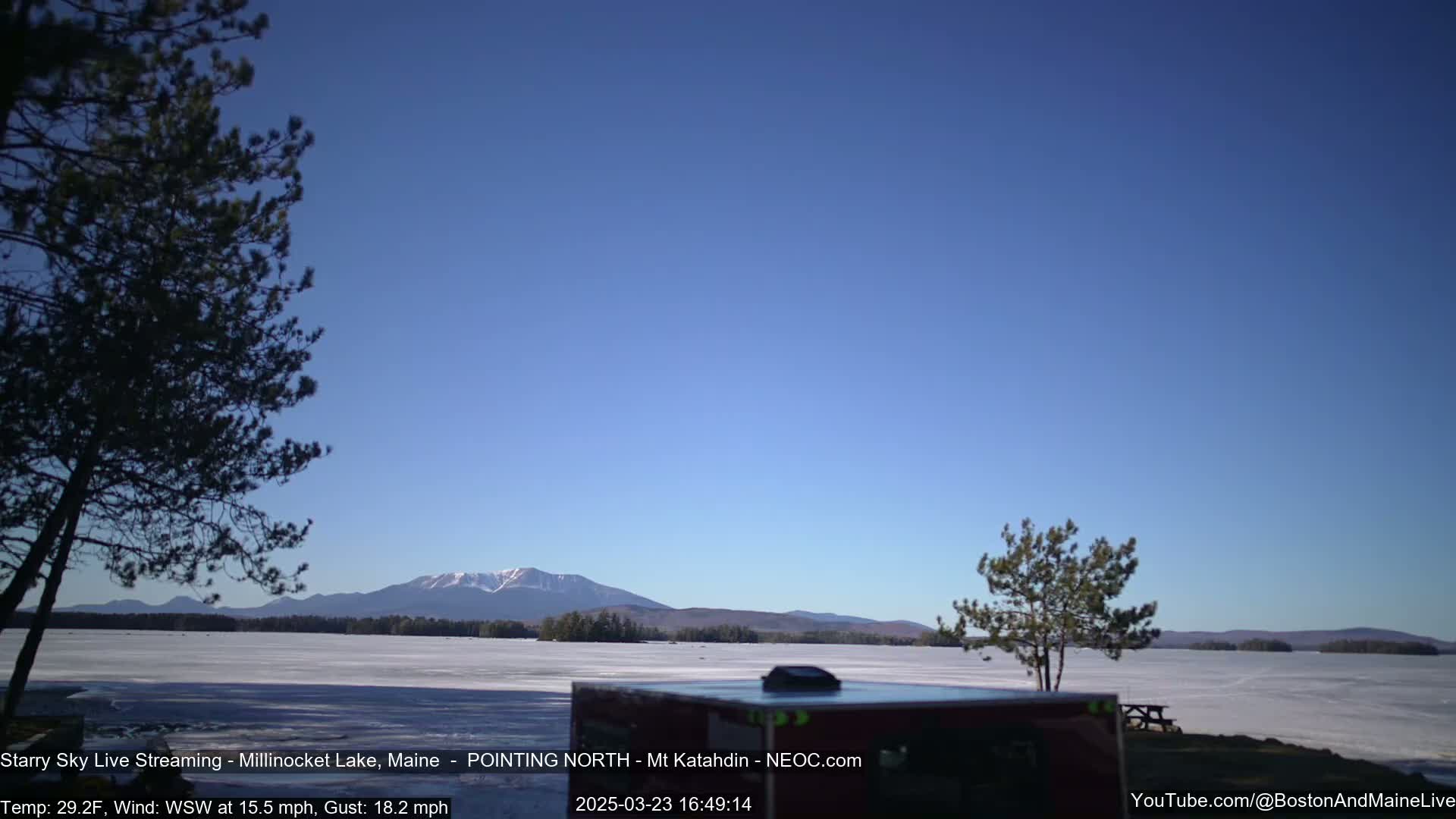 A frozen lake stretches out before a mountain range under a clear blue sky, with a few trees in the foreground and a vehicle partially visible.