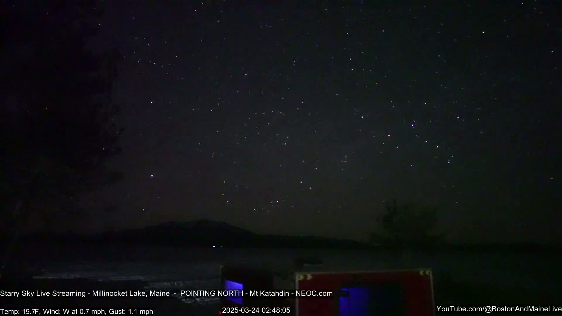A nighttime view of a star-filled sky over a dark lake with a distant mountain range, under calm, cool conditions.