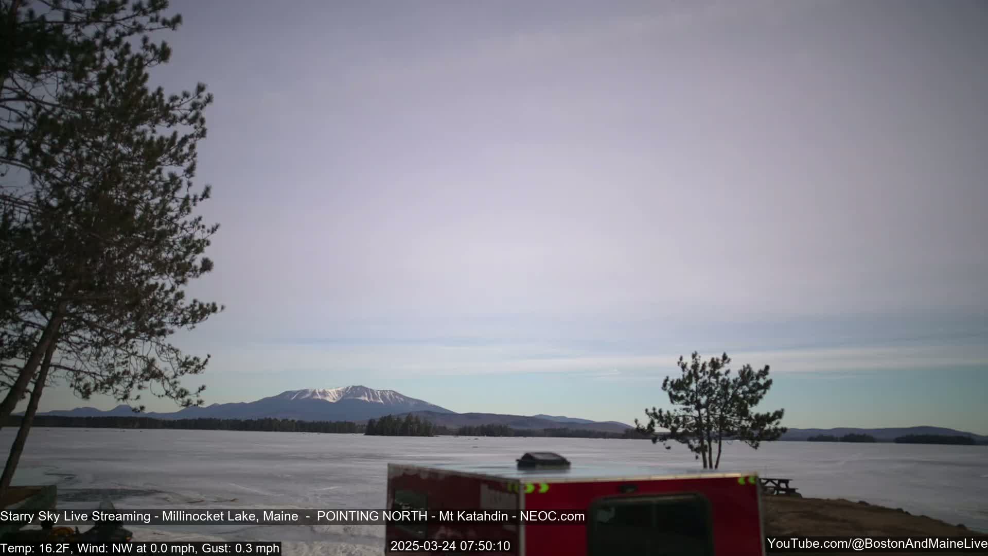 A snow-covered lake stretches to a mountain range under a pale sky, with a few trees in the foreground.