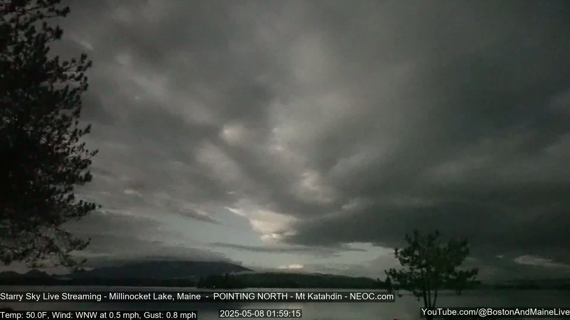 A mostly cloudy, dark sky over a calm body of water, with a few trees in the foreground and mountains in the distance.