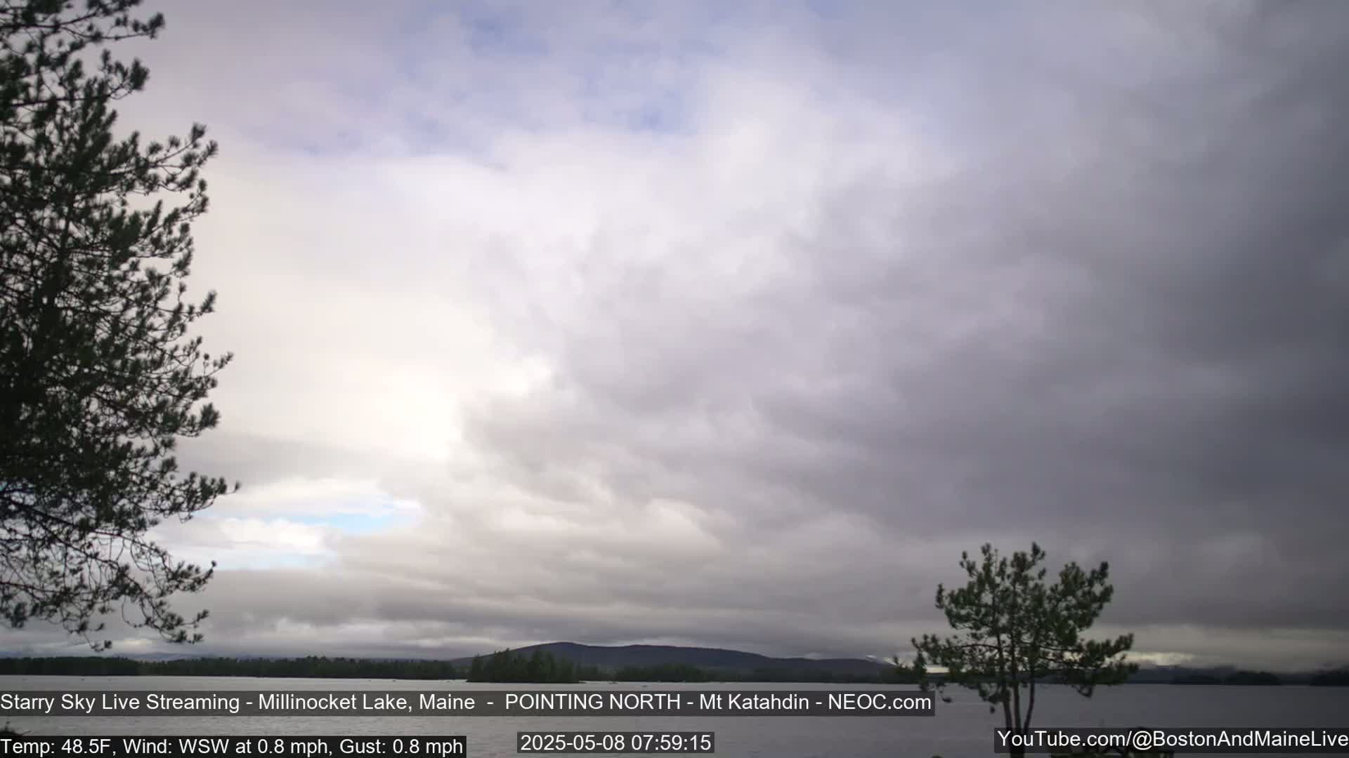 A mostly cloudy sky over a calm lake with dark evergreen trees on either side of the frame and distant mountains.