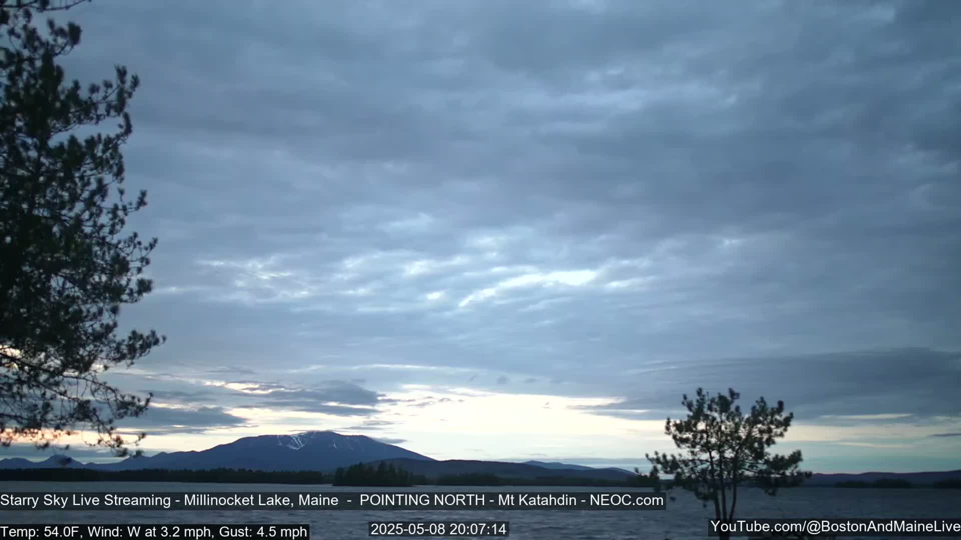 A calm lake is visible under a mostly cloudy sky, with a mountain range in the distance.