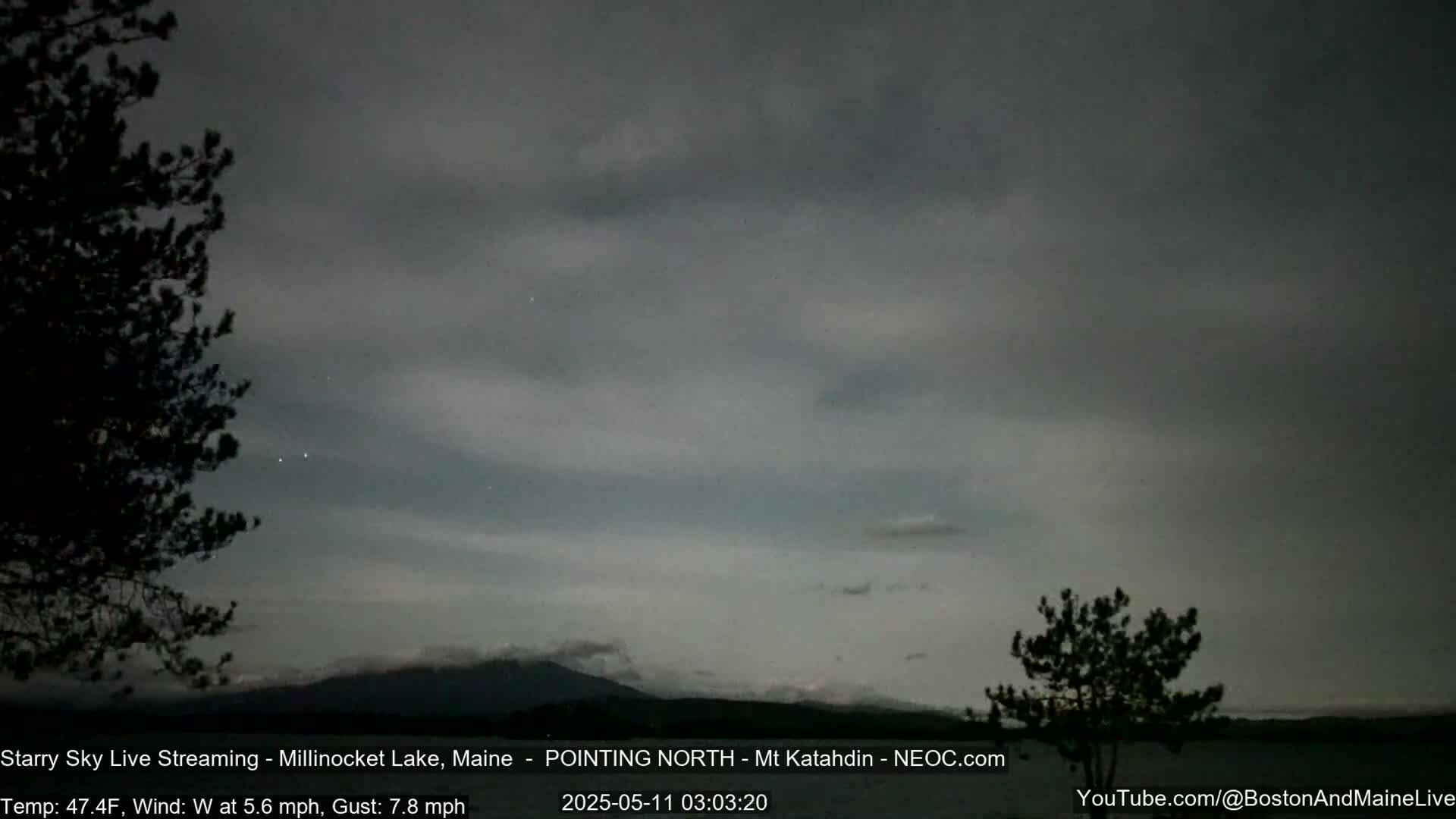 A dark, overcast night sky over a lake, with a mountain in the distance and silhouetted trees in the foreground, under a 47.4°F temperature with a 5.6 mph westerly wind gusting to 7.8 mph.