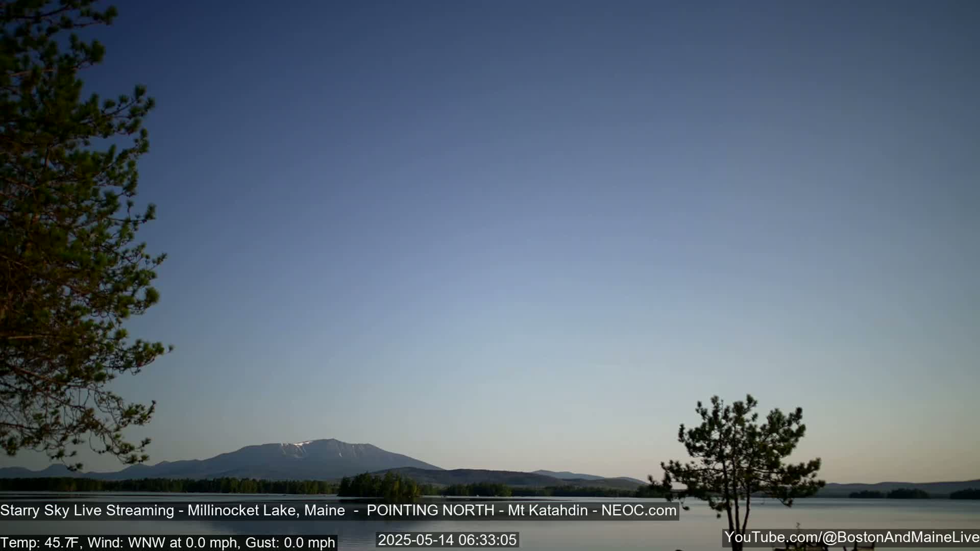 A calm lake is seen with a mountain range in the background under a clear, twilight sky.
