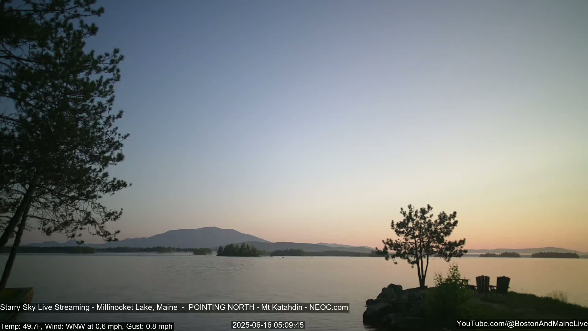 A calm lake at sunrise, with a mountain range in the background under a clear, pale sky.