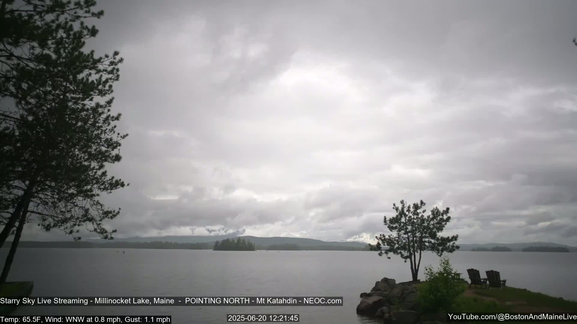 A calm lake under an overcast sky is viewed from a rocky shoreline with two chairs and a small tree.