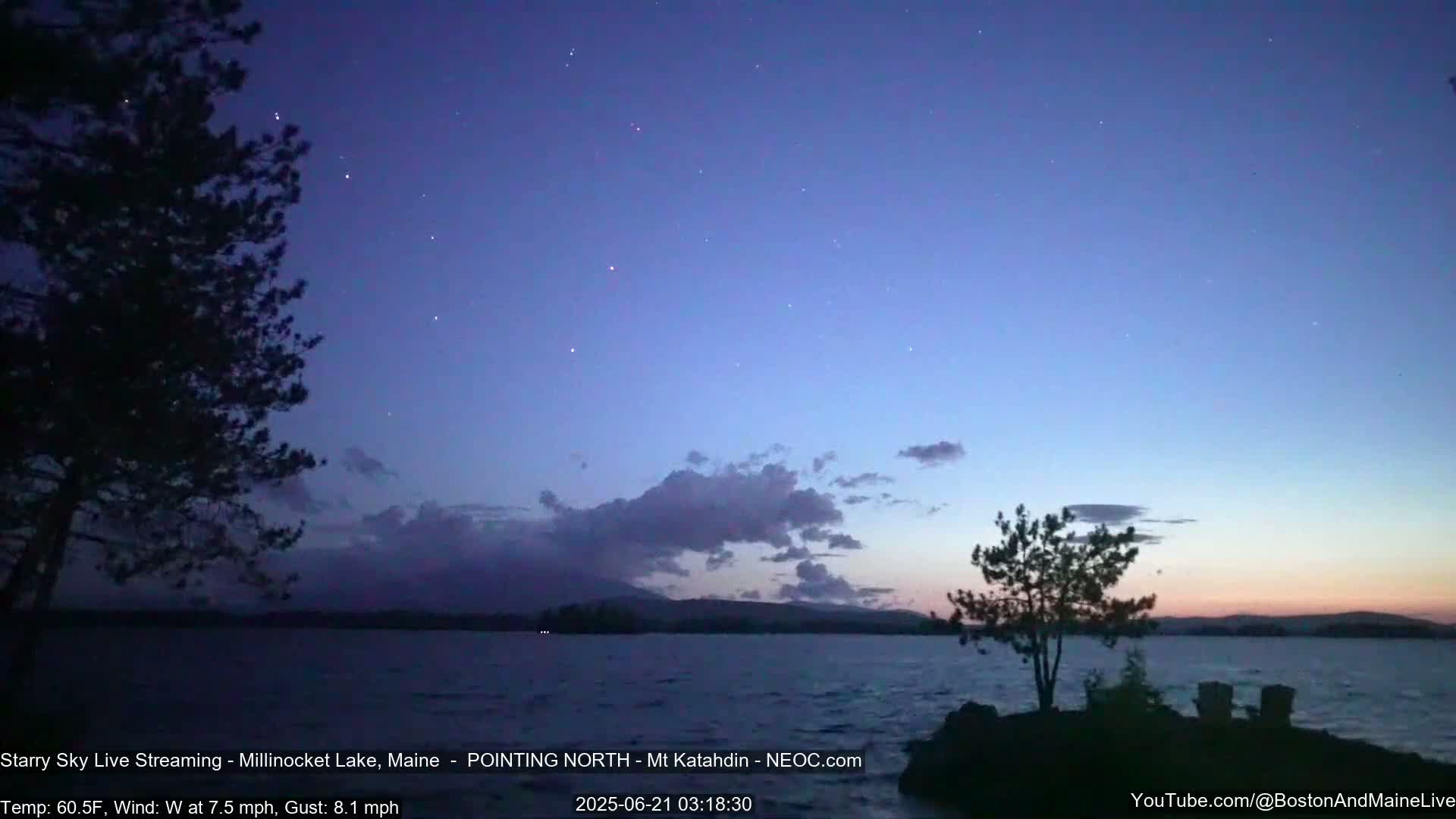 A calm lake at twilight under a starry sky with a few clouds and a gentle wind, silhouetted by trees and a distant mountain range.