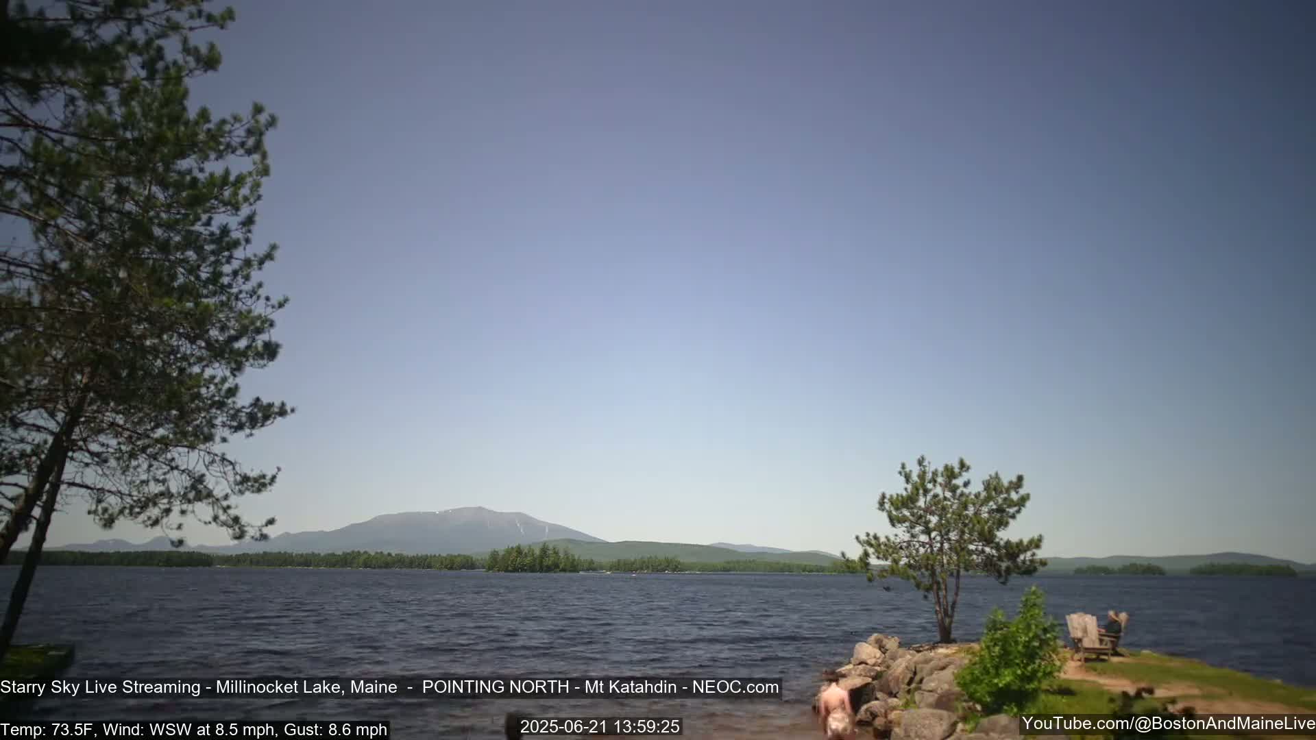 A calm lake with a distant mountain range under a clear, light-blue sky, with a slight wind.