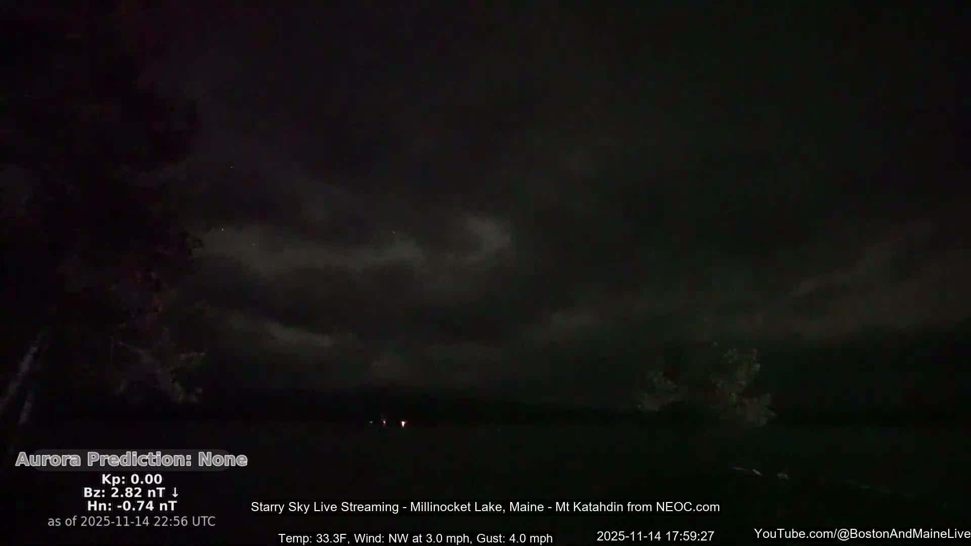 A dark, cloudy night sky hangs over a still body of water, with silhouetted trees framing the sides and a few faint lights visible in the distance.