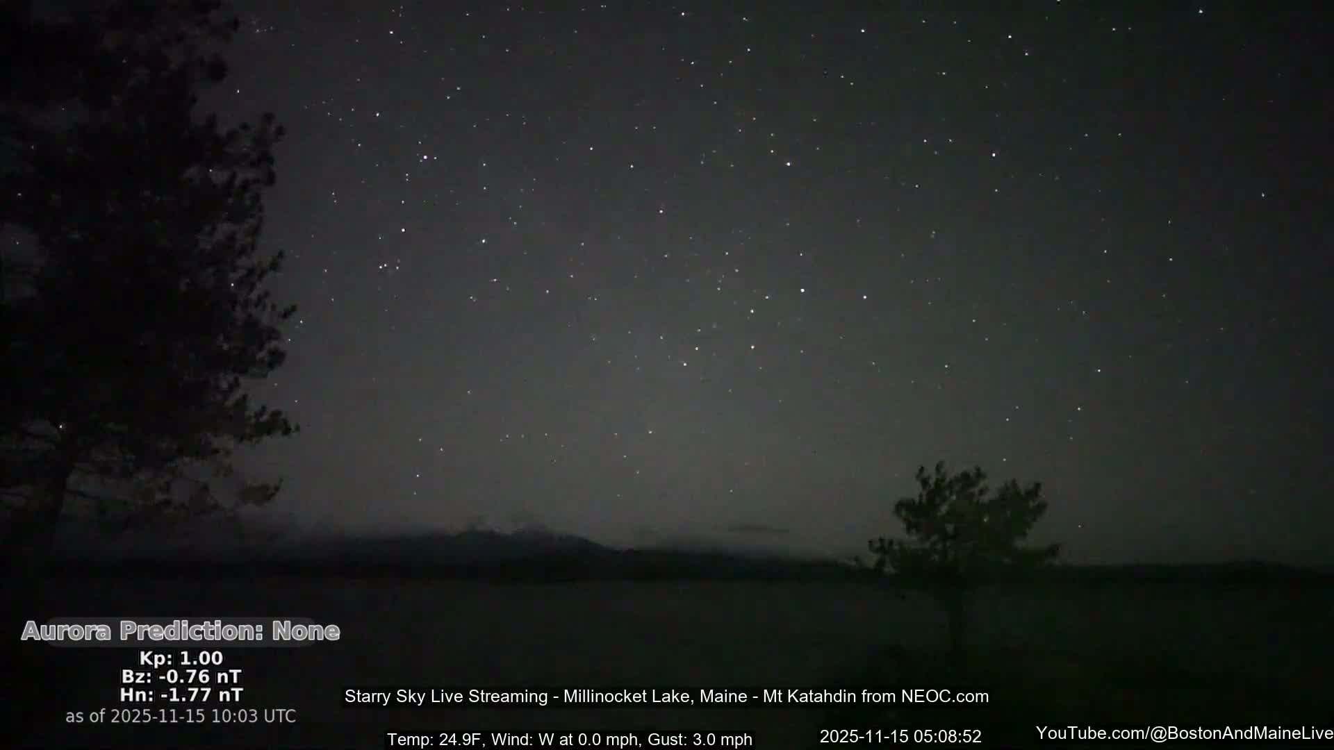 A clear, cold, and calm starry night sky stretches over a dark body of water, flanked by silhouetted trees and distant mountains.
