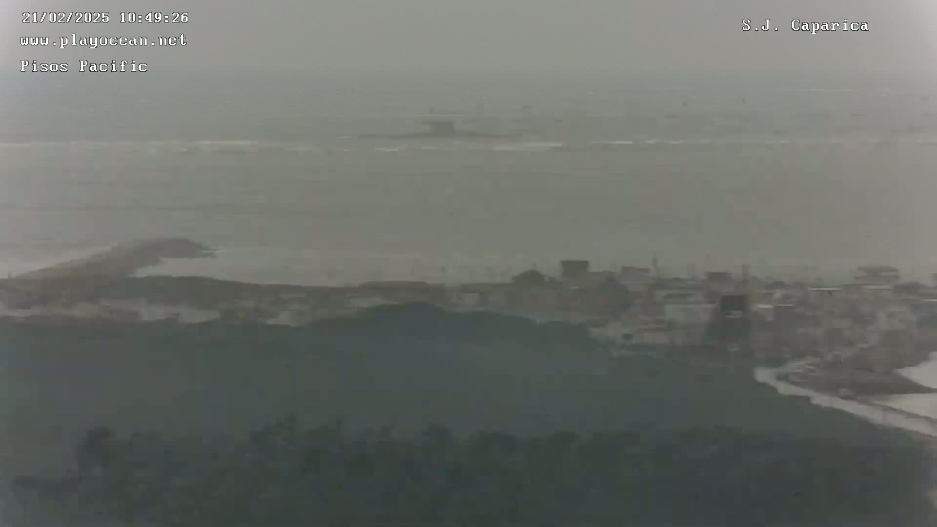 A hazy, overcast view shows a coastal town nestled between a body of water and rolling hills.