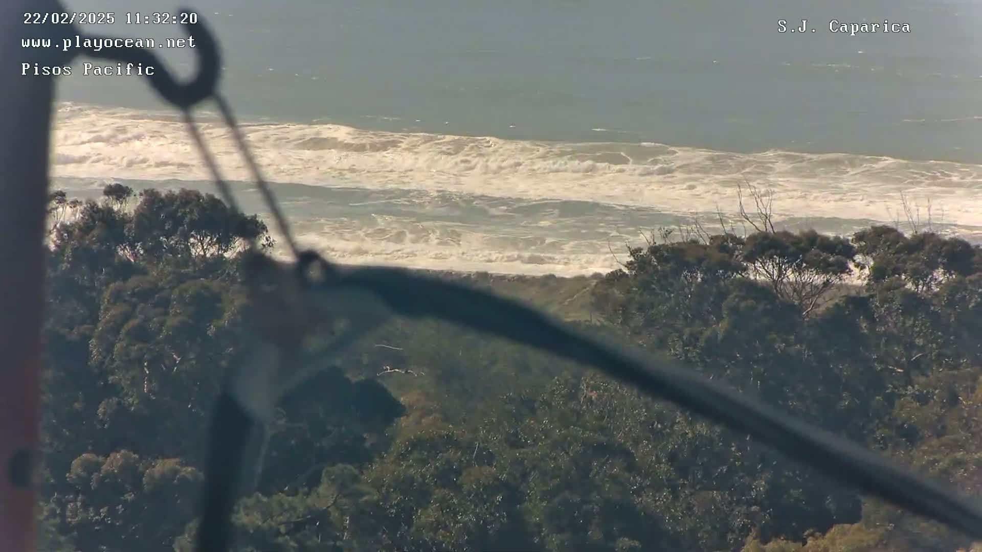 A view from above shows a turbulent ocean with white-capped waves crashing against a sandy shore, partially obscured by a dark object and a foreground of dark green trees under a mostly clear sky.