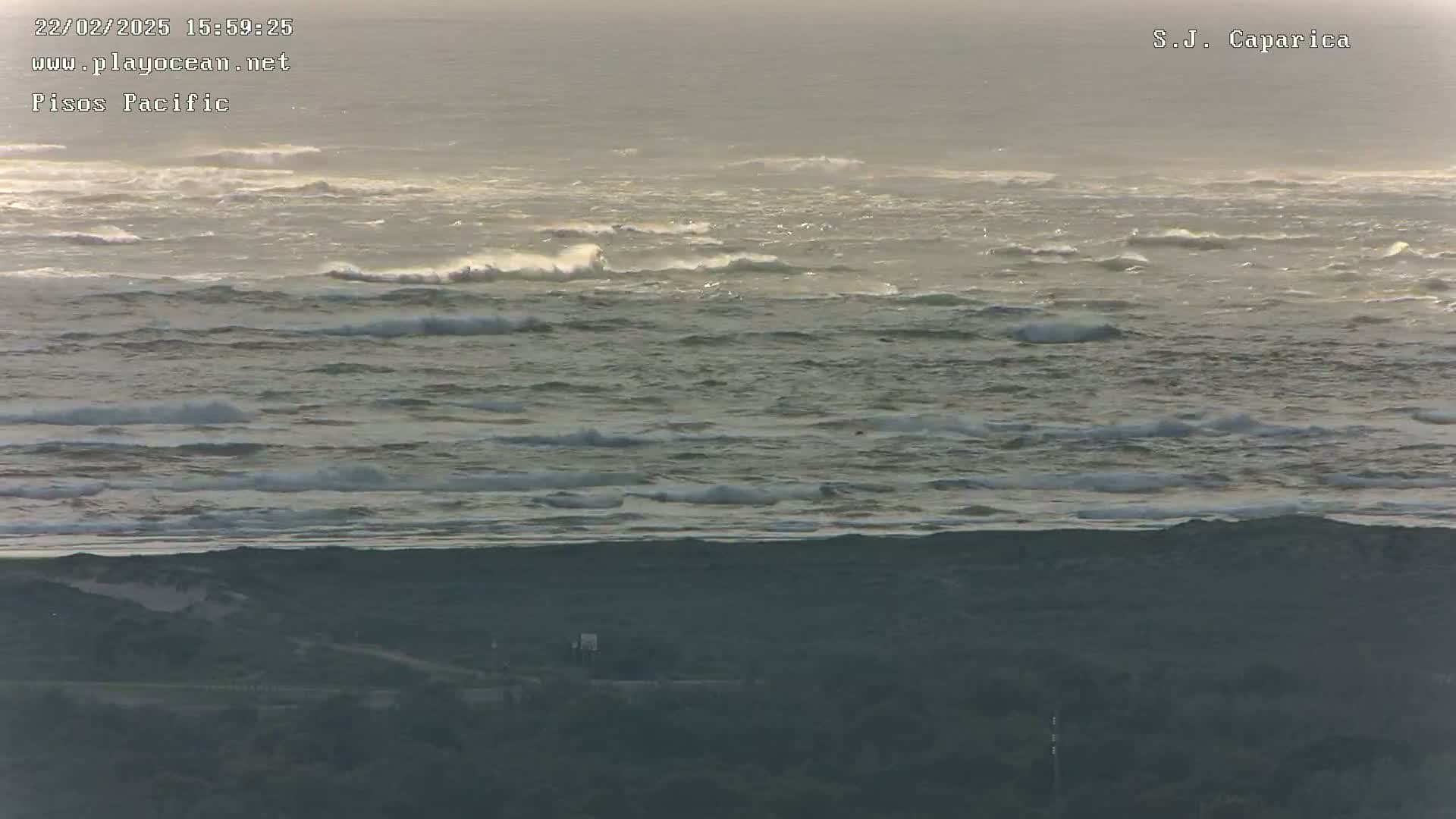 A hazy, overcast day shows a wide expanse of ocean with moderate waves breaking, and a dark shoreline in the foreground.