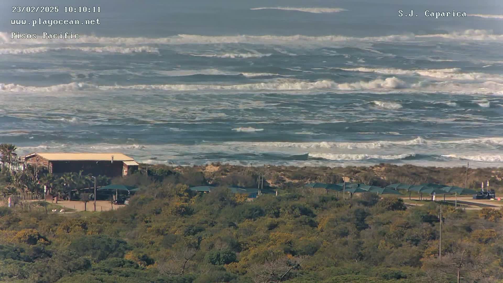 A building sits near a sandy shore with choppy waves and whitecaps under a mostly clear sky.
