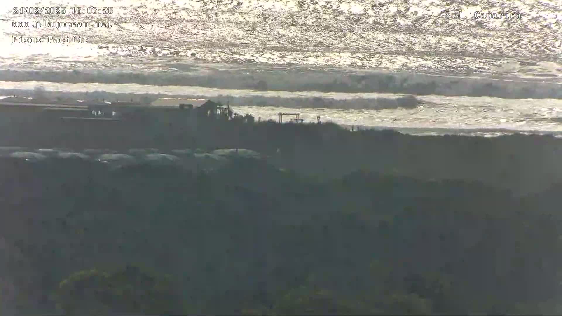 A distant view shows a building near the ocean under a bright, sunny sky, with waves gently rolling in.