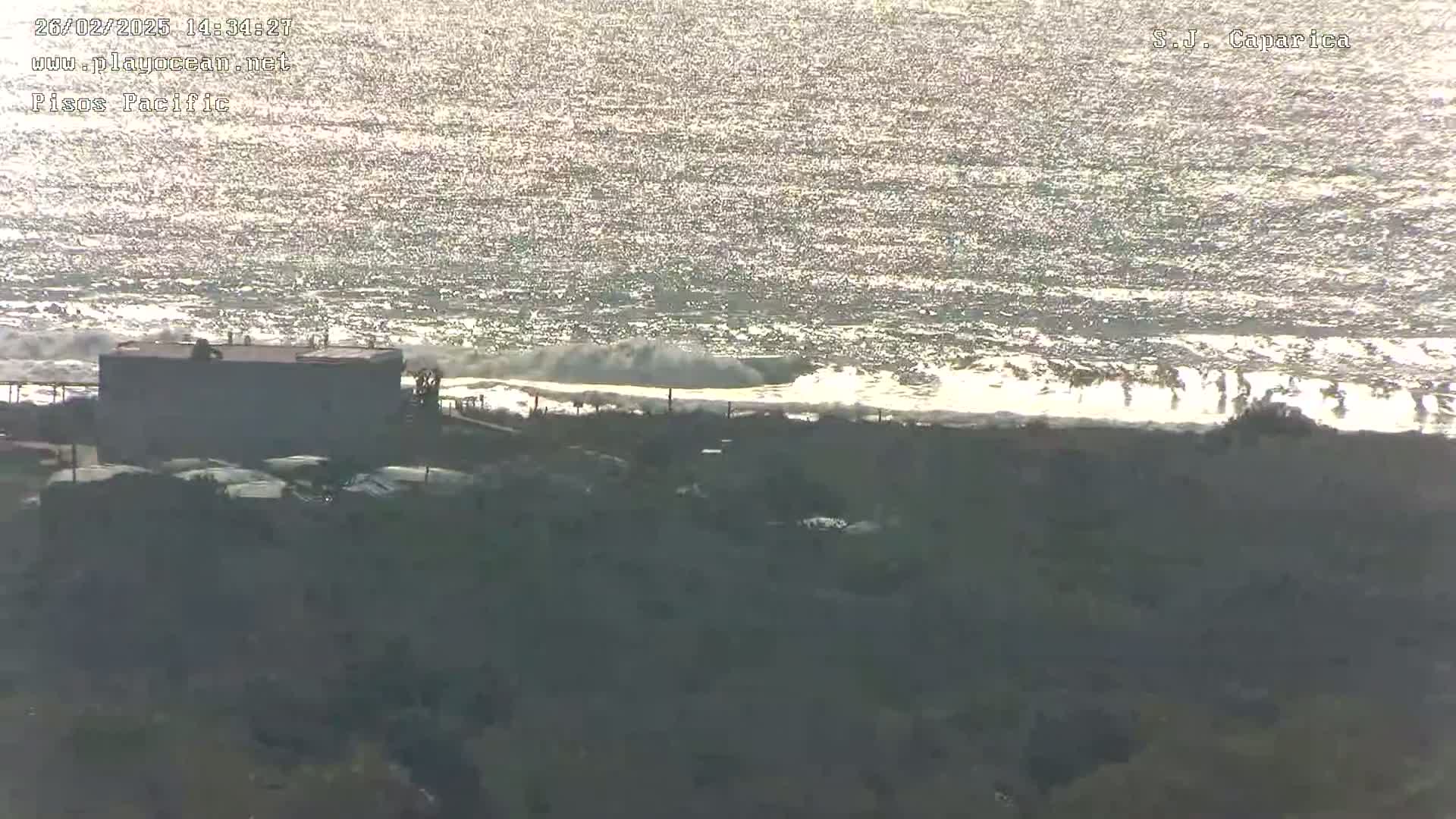 A mostly sunny day shows a glittering ocean with waves breaking on a shore where several buildings and vehicles are visible from an elevated vantage point.