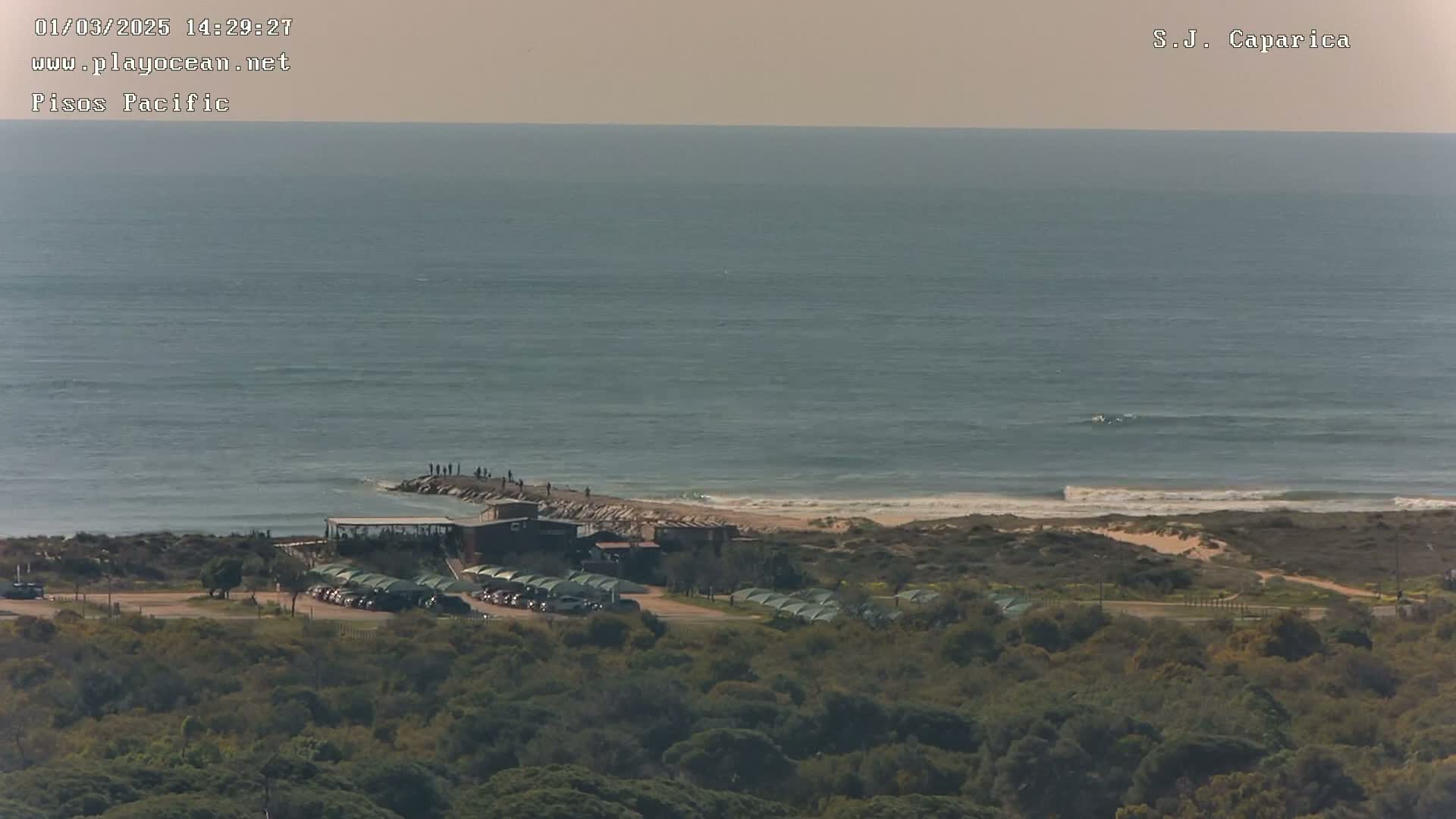 A beach scene shows a calm ocean with a few people on a jetty, a small building, parked cars, and green vegetation in the foreground under a hazy sky.