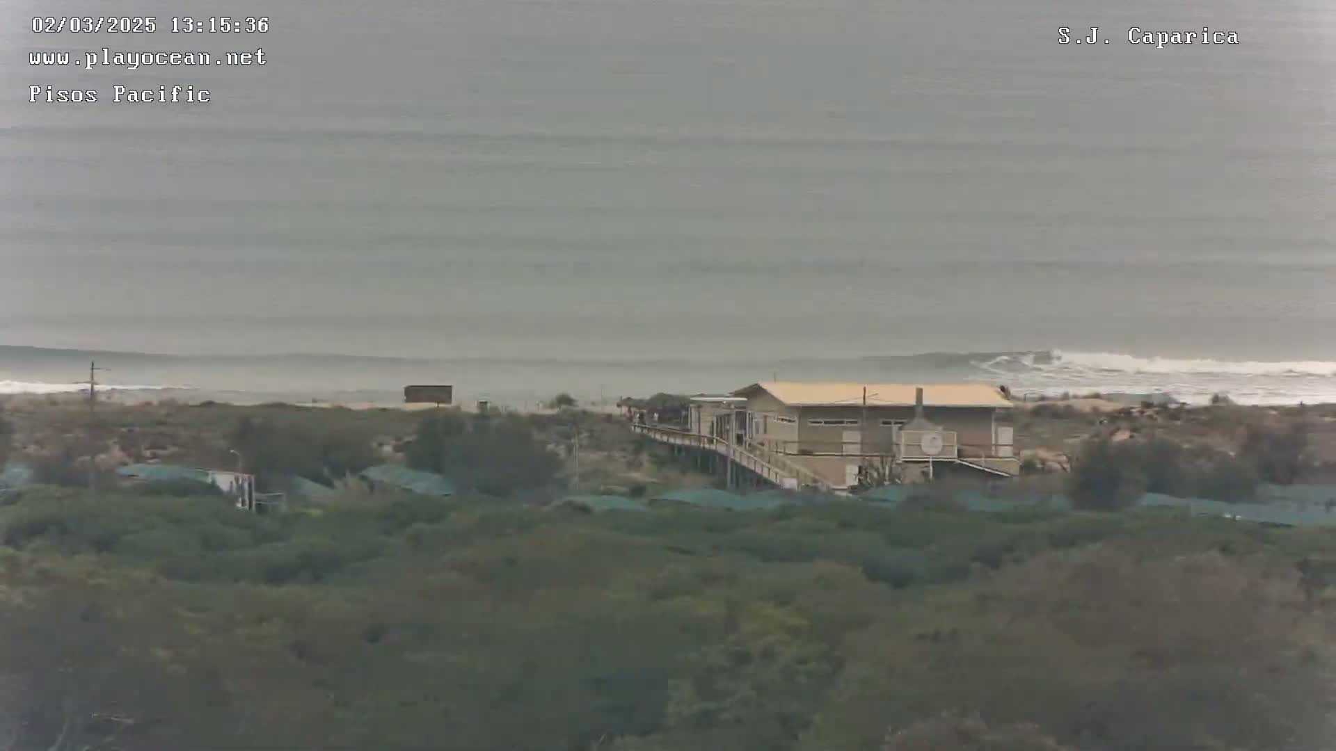 A long, gray, overcast beach scene shows a building near the ocean with low-lying vegetation in the foreground.
