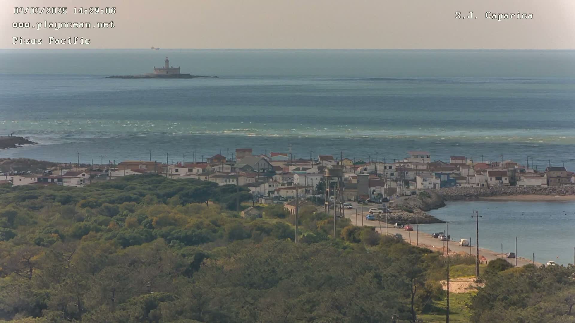 A coastal town with a lighthouse visible in the distance across a calm, slightly wavy ocean under a hazy sky.