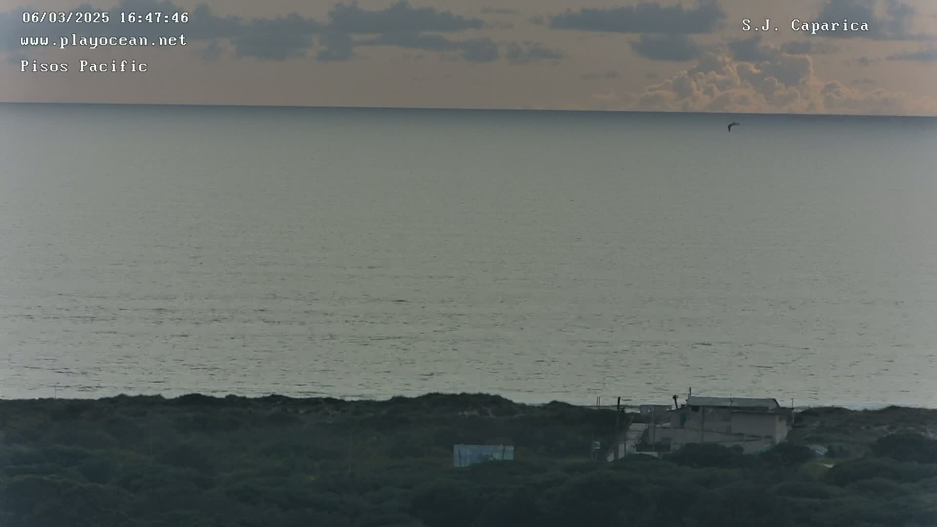A calm ocean under a mostly cloudy sky is visible from a high vantage point overlooking a low-lying structure and vegetation.