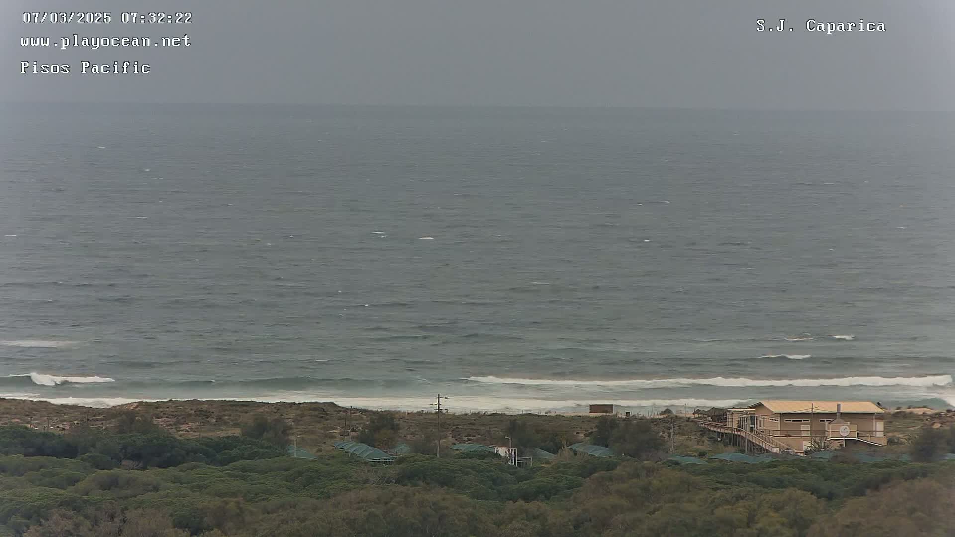A gray, overcast day shows a choppy ocean meeting a sandy shore lined with low-lying vegetation and a single-story building.