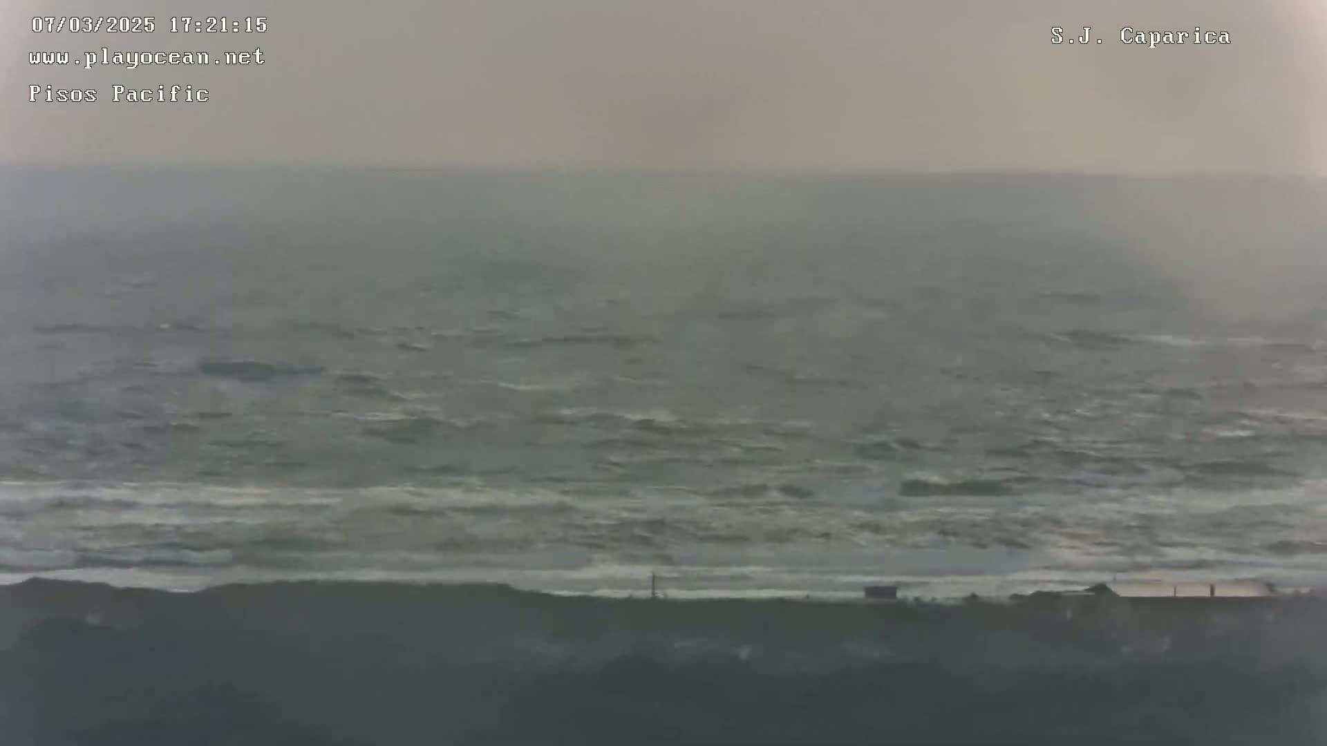 A hazy, overcast day reveals a choppy ocean meeting a dark shoreline with a few structures visible in the distance.