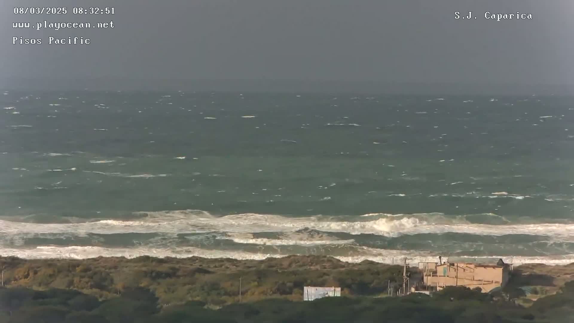 A stormy ocean with choppy waves crashes against a shore where low-lying vegetation and a small building are visible.
