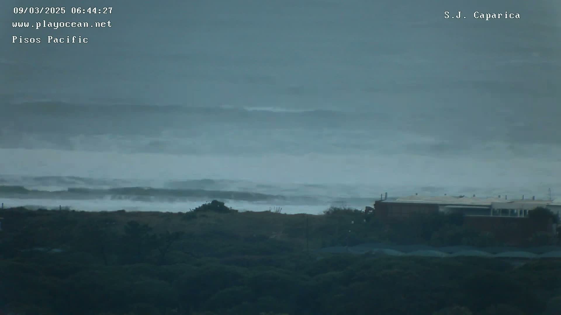 A misty, overcast day reveals distant, rolling waves crashing on a shore, behind which sits a low-lying building nestled amongst dark green vegetation.