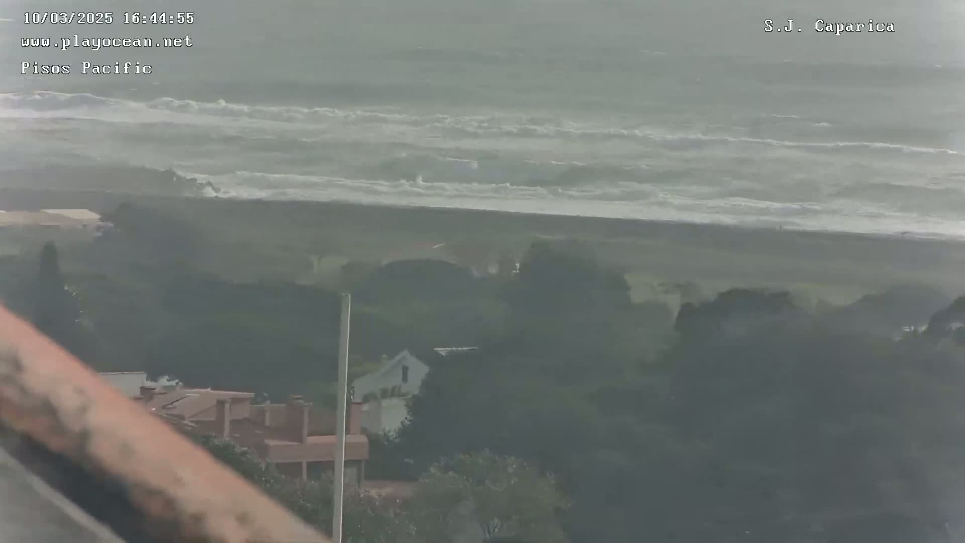 A hazy view shows a beach with choppy waves under an overcast sky, overlooking a coastal residential area.