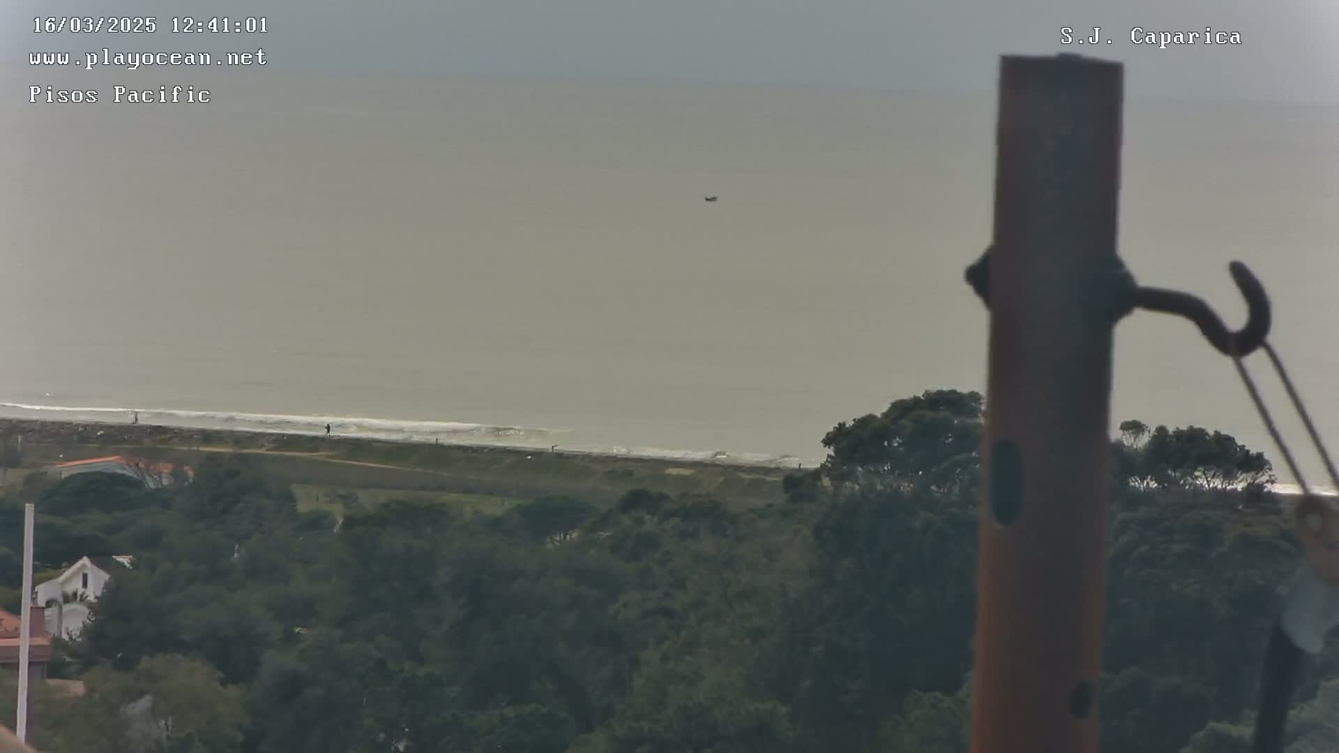 A distant view of a coastline with a calm, gray sea under an overcast sky, seen from behind a rusty metal post.
