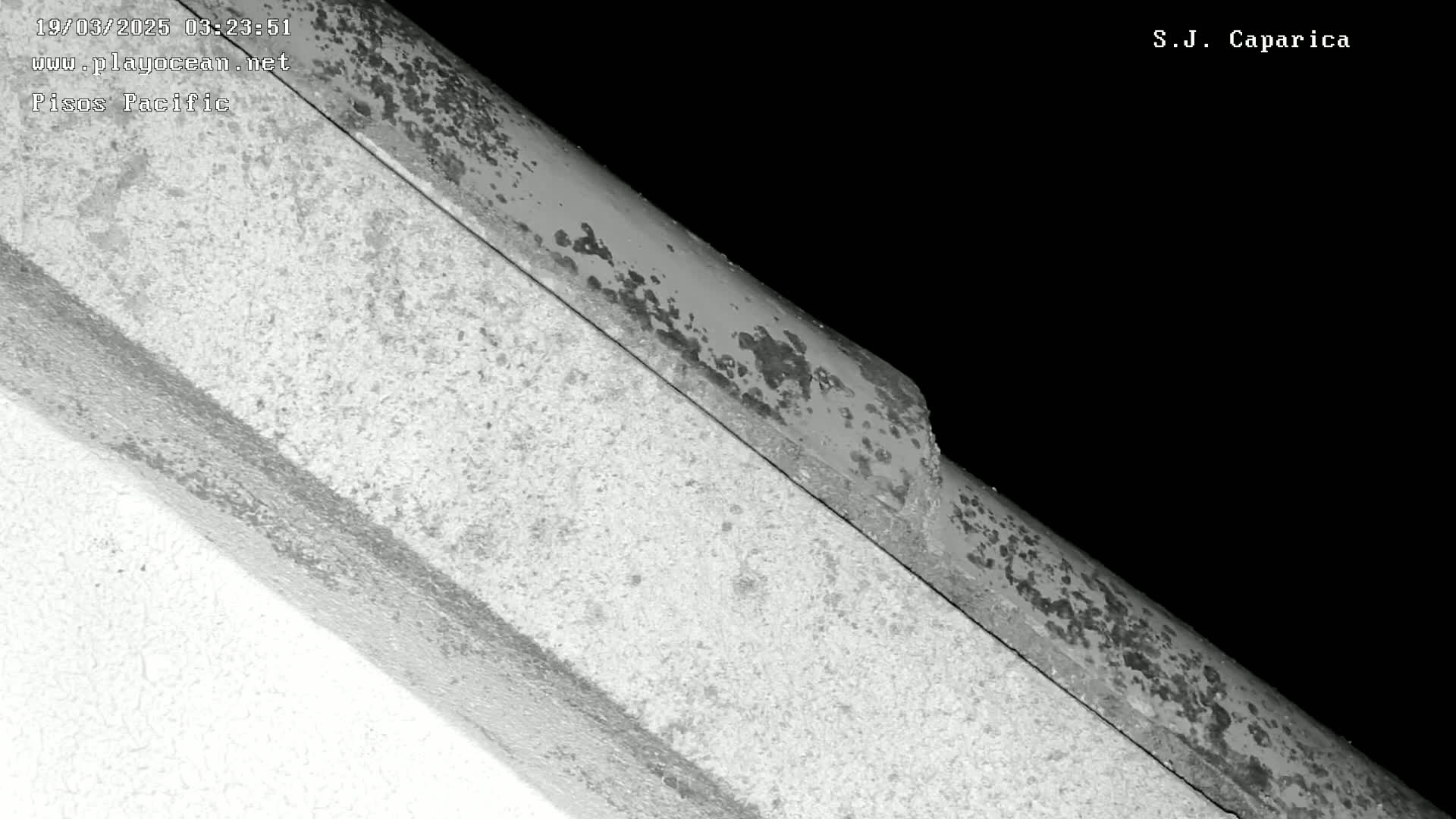 A close-up, nighttime grayscale shot shows a weathered, rusty metal piece angled against a lighter, textured surface.