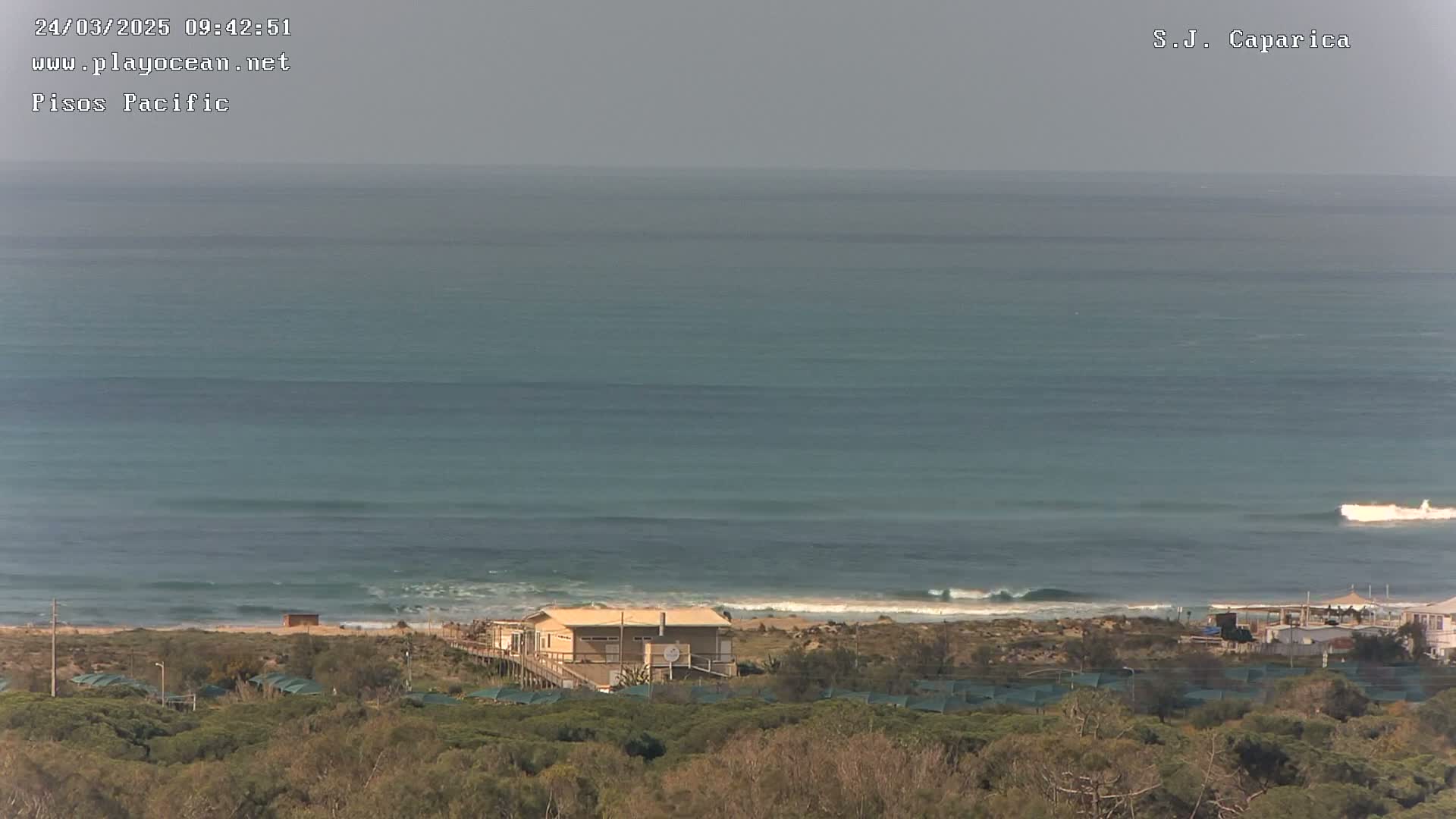 A coastal scene on an overcast day shows a calm ocean with small waves breaking near a sandy beach, behind which sits a building and low-lying vegetation.