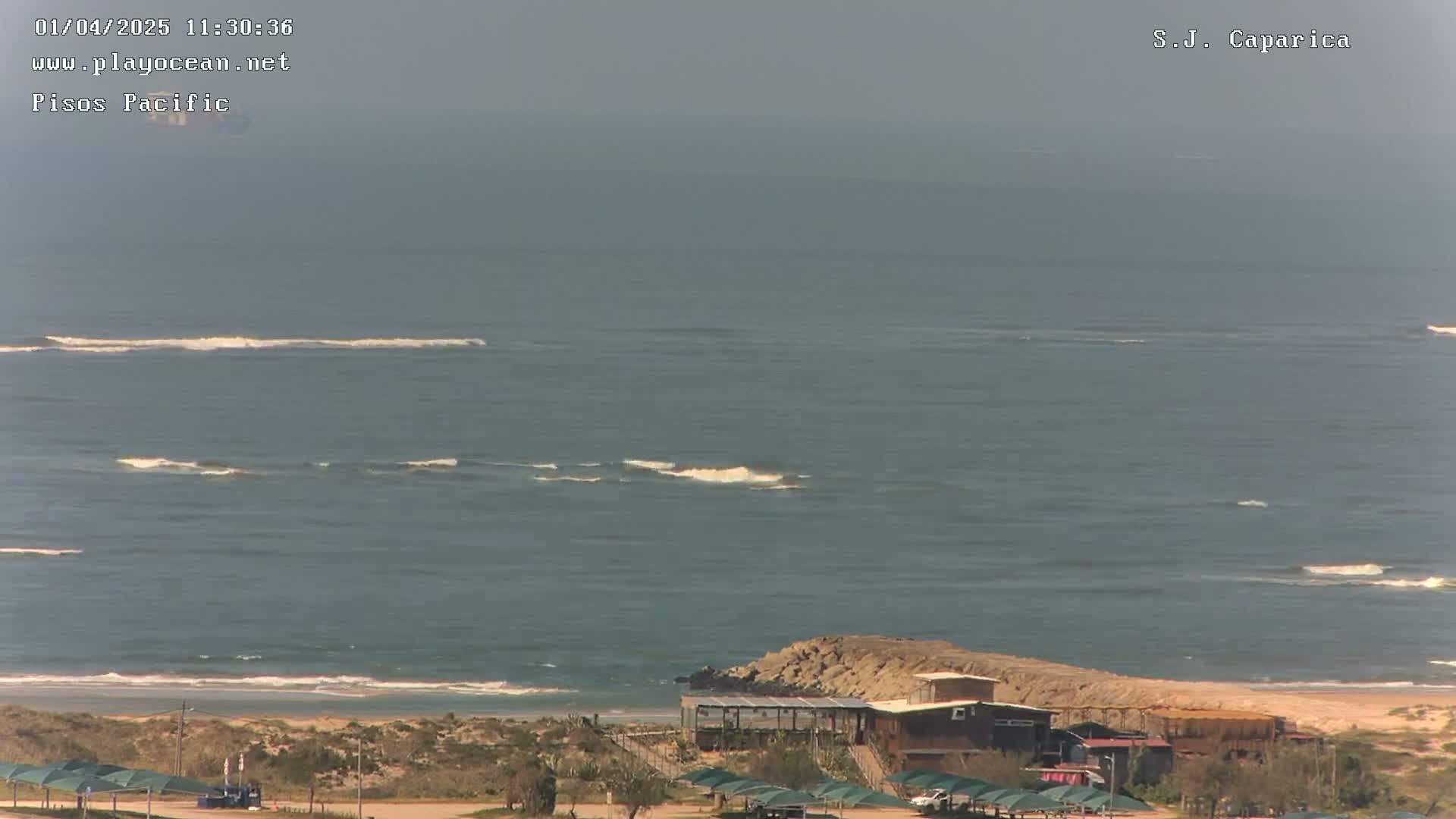 An ocean with small waves under a hazy sky, viewed from a high vantage point overlooking a sandy beach and a few buildings.
