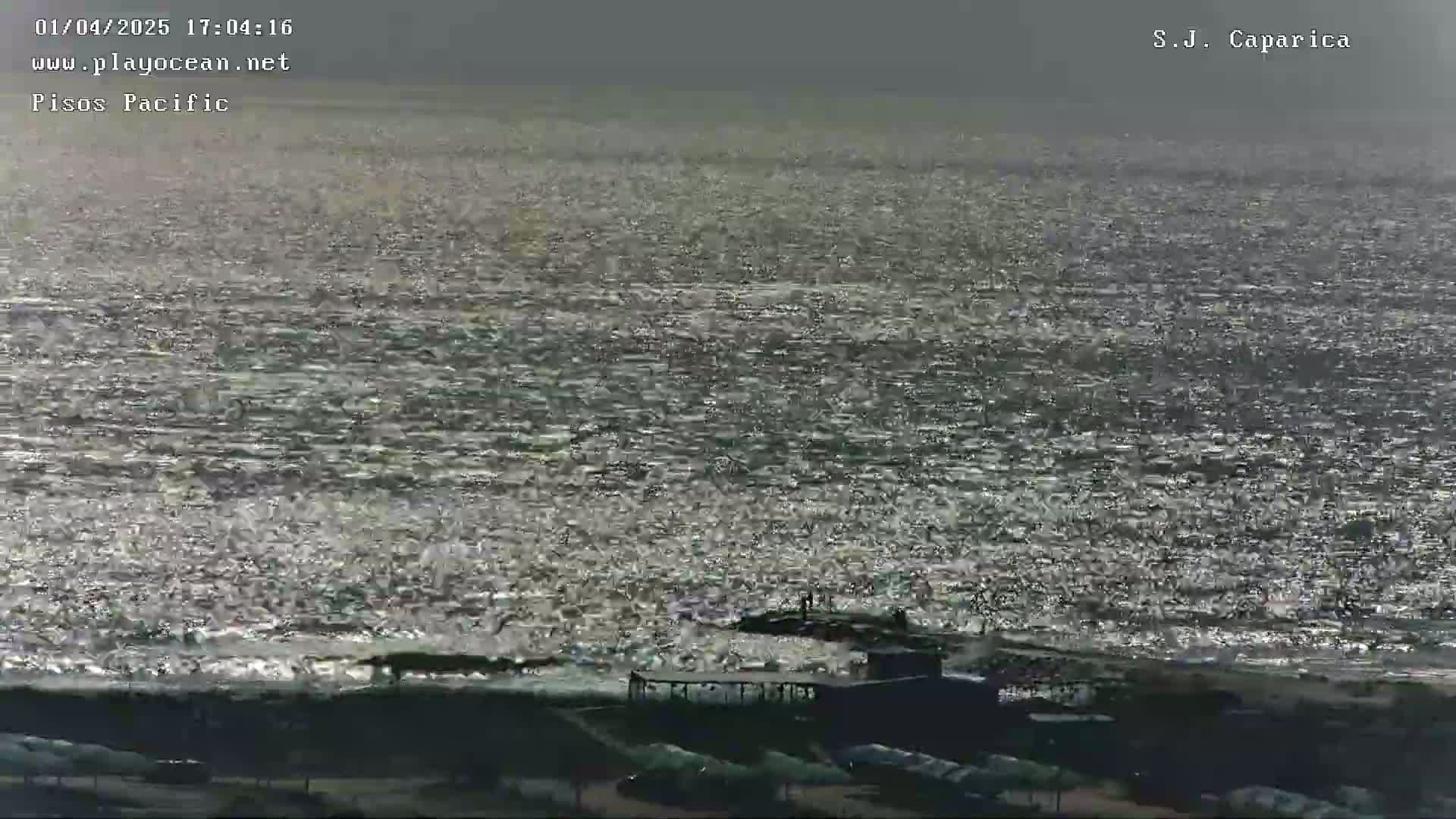 A hazy, sun-dappled ocean meets a dark shoreline with a few structures near the water's edge.