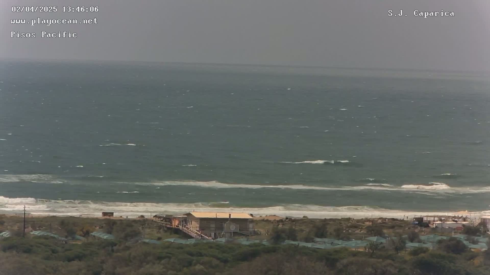 A slightly overcast day shows a choppy ocean with small waves breaking on a sandy beach, behind which sits a cluster of low-lying buildings surrounded by vegetation.