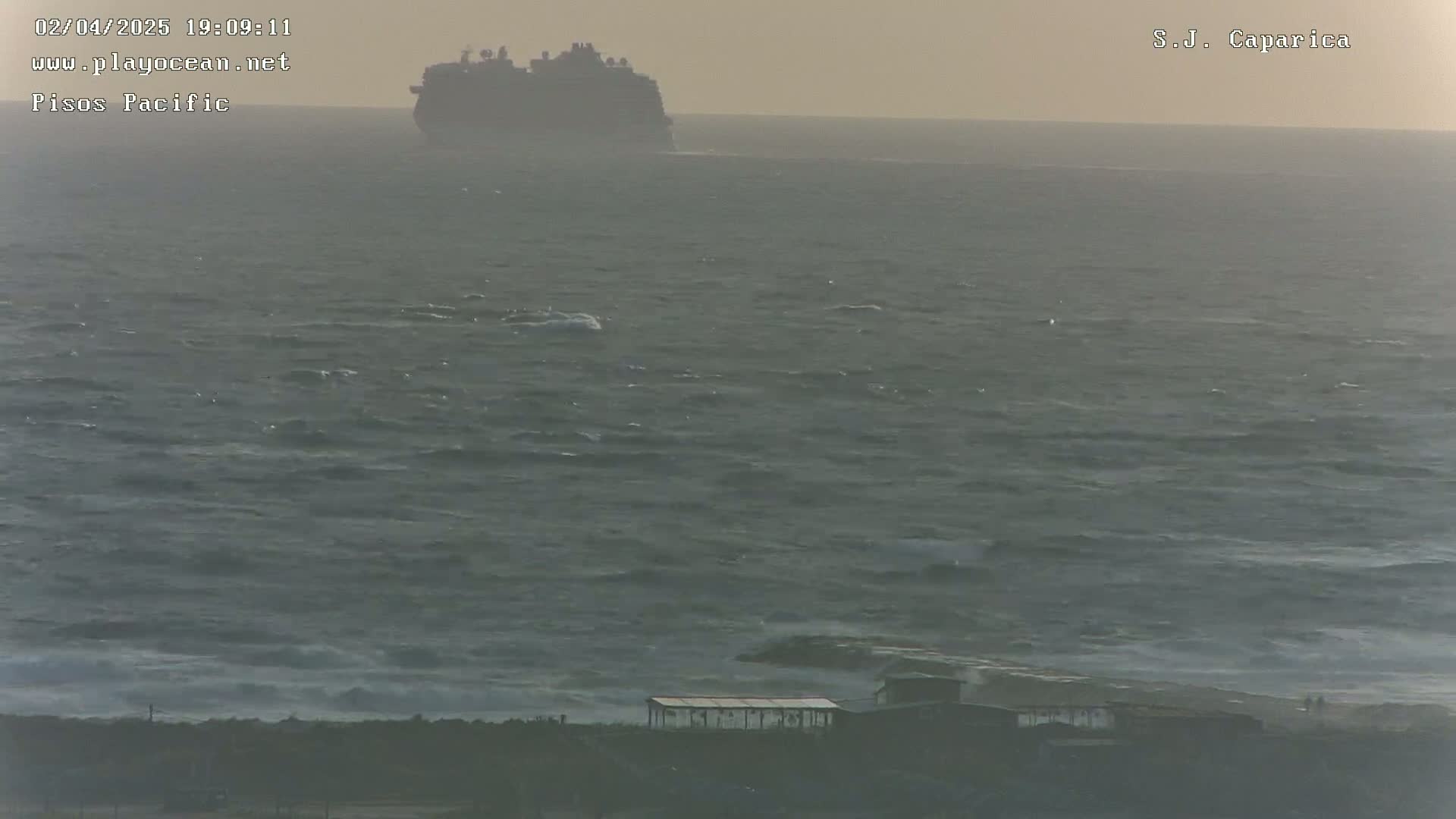 A large cruise ship is visible on the horizon over a choppy, gray sea, with a small structure and some people visible on the shore in the foreground.