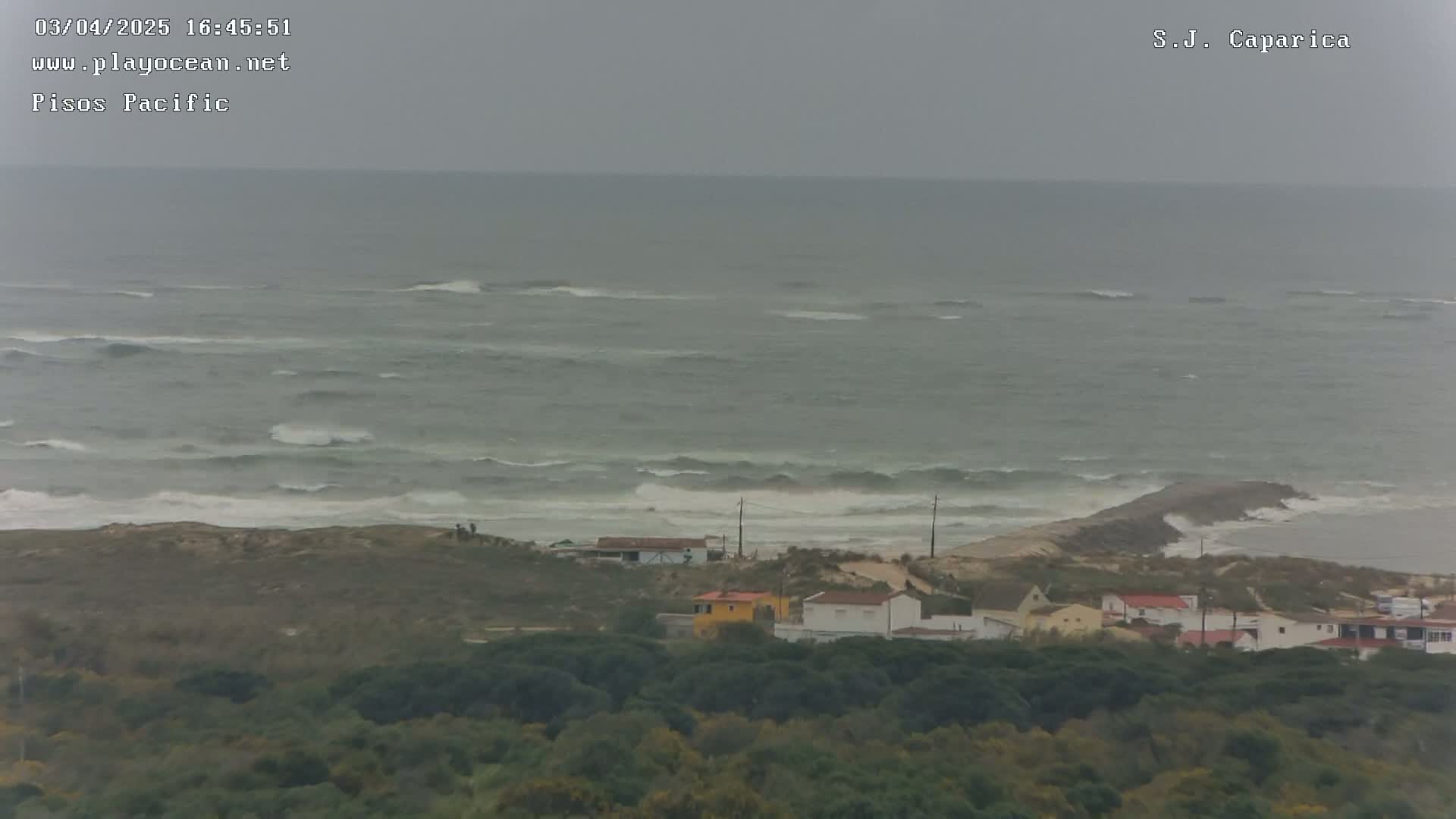 A view of a gray, choppy ocean with waves breaking near a coastline where several houses are nestled among green vegetation.