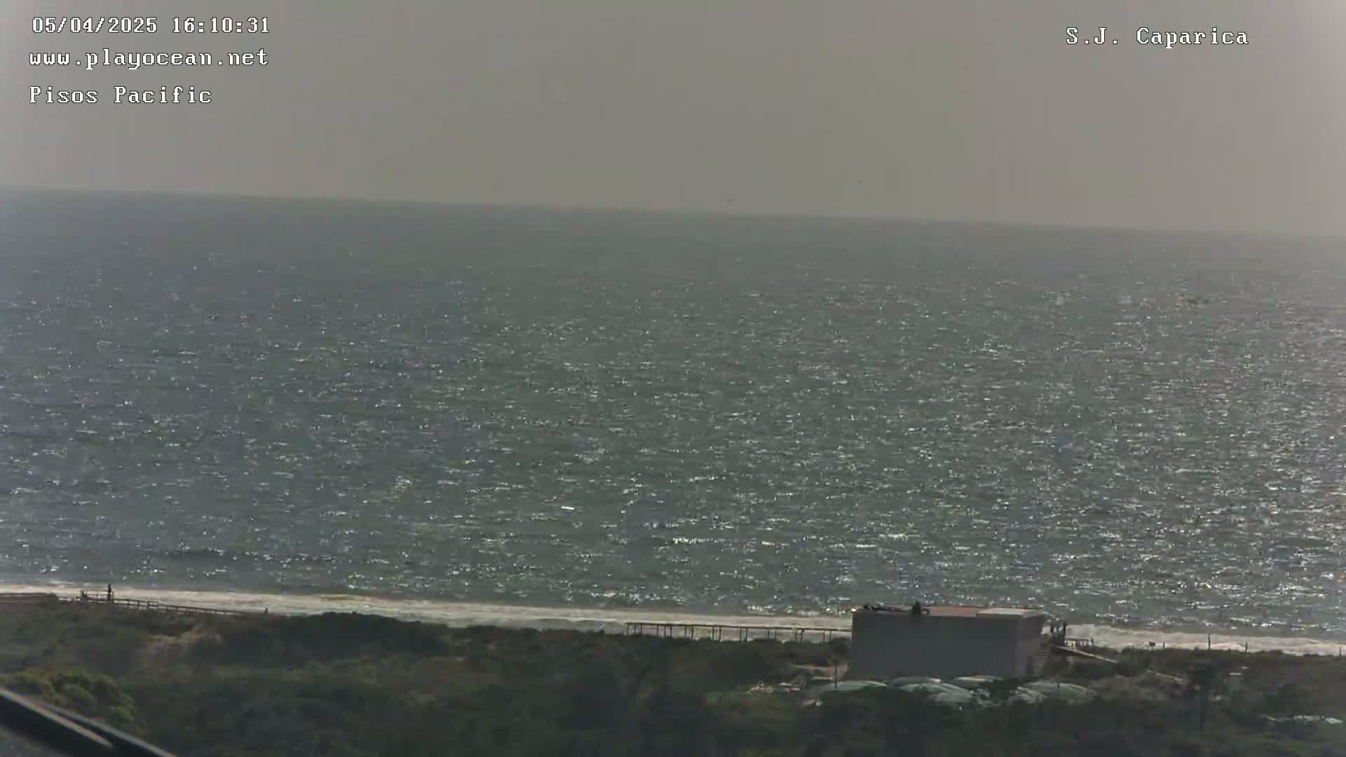 A slightly hazy, sunny day reveals a wavy ocean meeting a sandy shore, behind which sits low-lying vegetation and a small building.