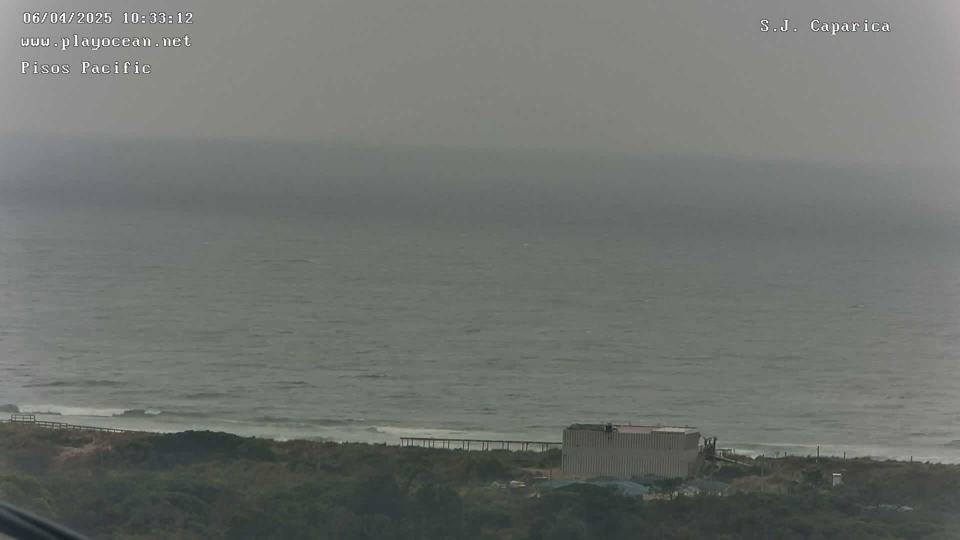 A hazy, overcast day shows a view of a grey ocean meeting a shoreline with low-lying vegetation and a large light-grey building.