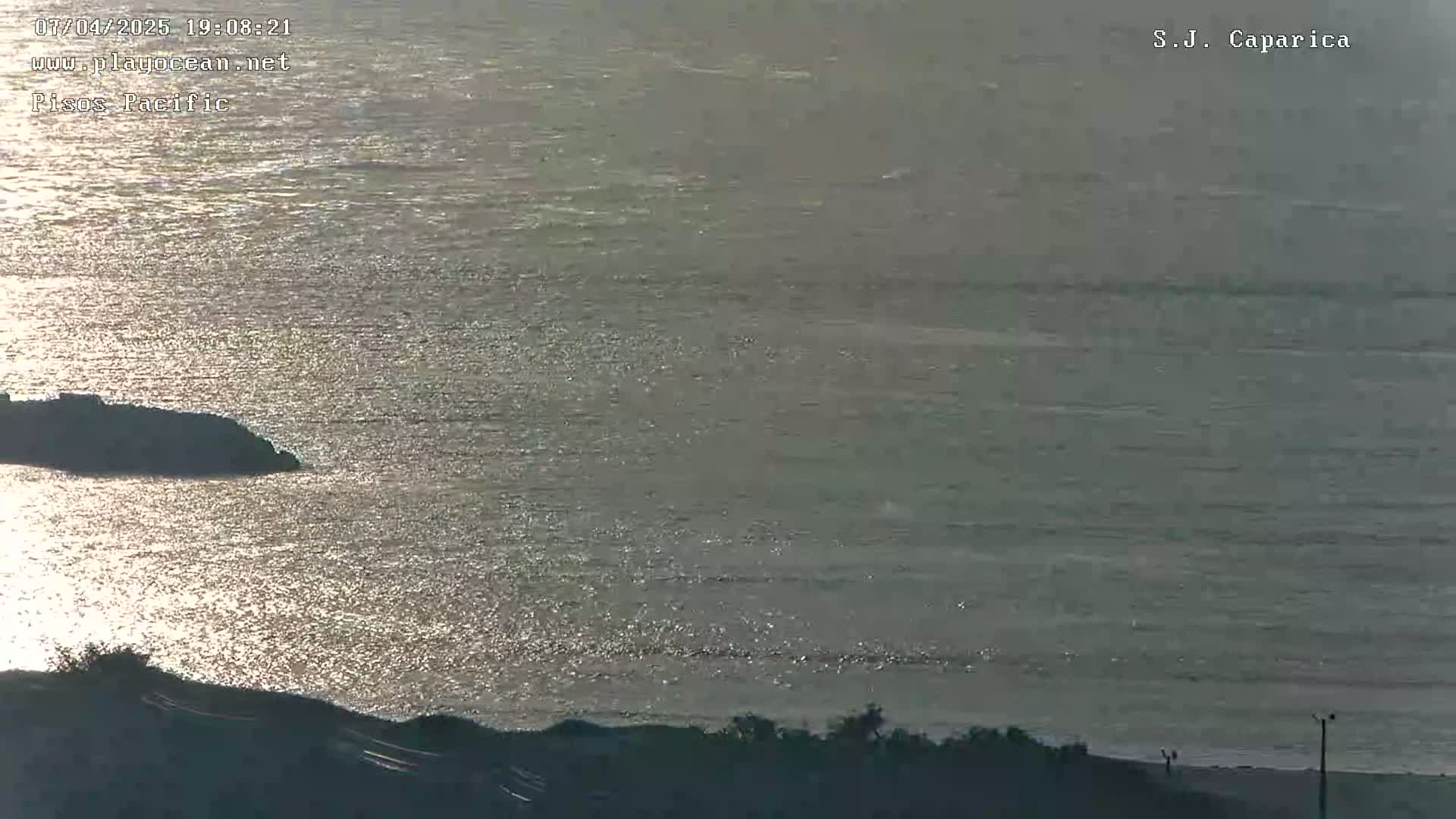 A sunlit ocean reflecting light, with a dark shoreline and land visible in the foreground.
